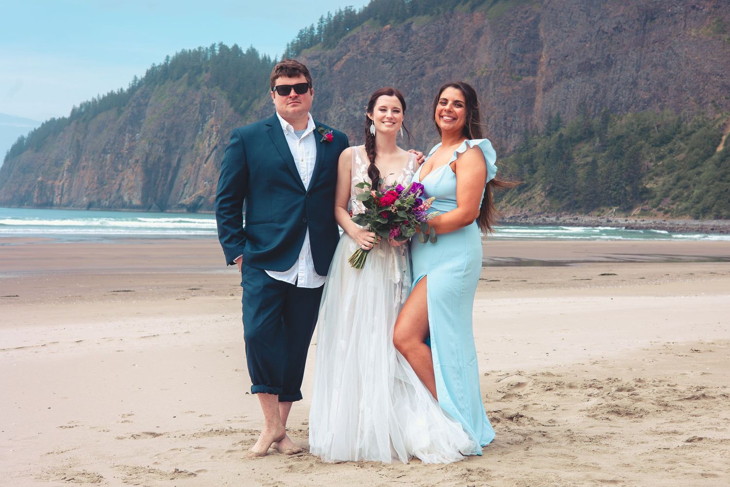 Groom in sunglasses stands with bride holding bouquet and bridesmaid on a sandy beach.