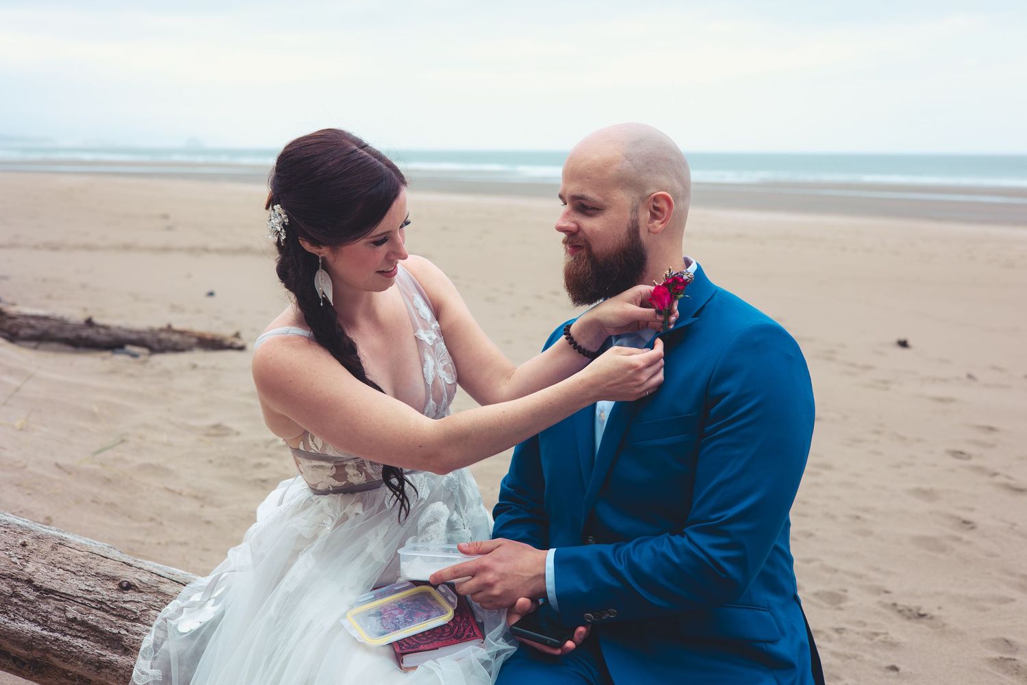 Bride pins boutonniere on groom in blue suit during beach wedding ceremony.