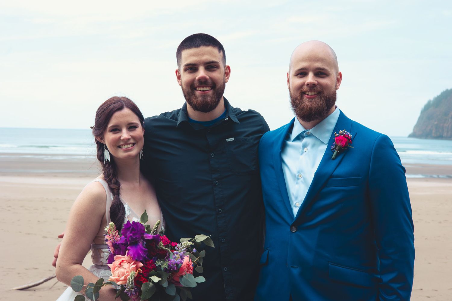 Bride and groom pose with a guest on a sandy beach, bride holding colorful bouquet.