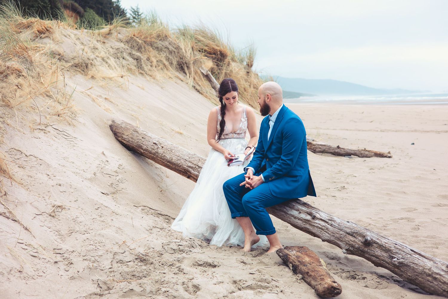 Bride and groom sit together on driftwood in beach sand dunes on their wedding day.