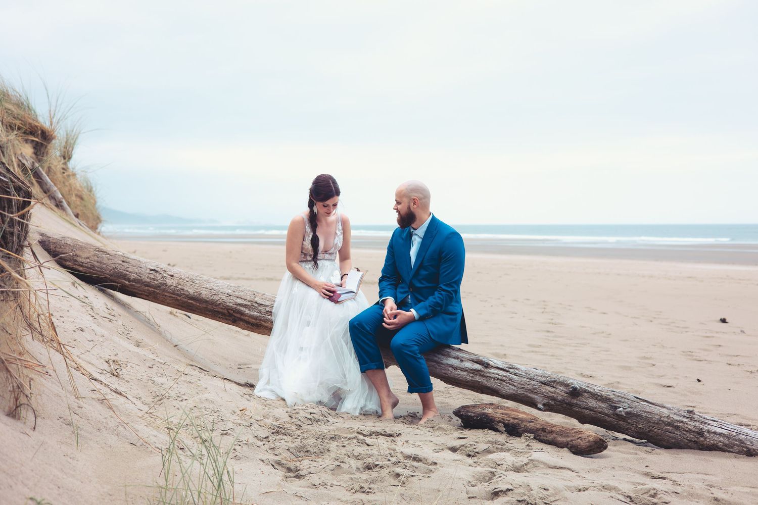 Bride reads from a small book while groom listens attentively, both seated on driftwood on a sandy beach.