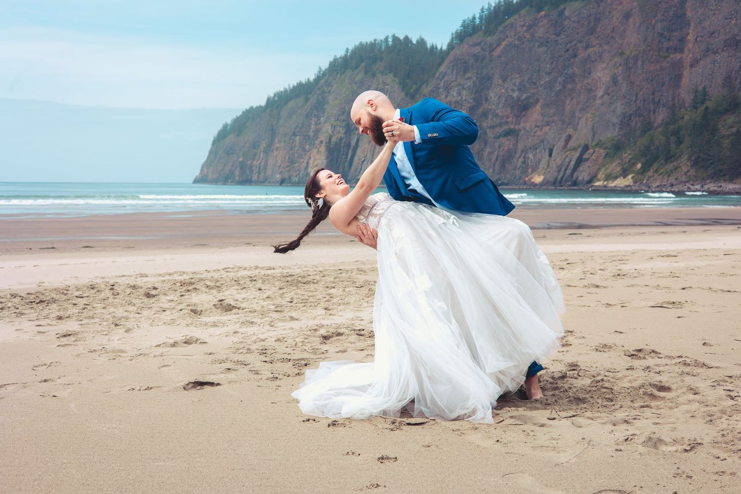 Groom dips bride dramatically on sandy beach, her white gown flowing, forested rocky cliffs in background.