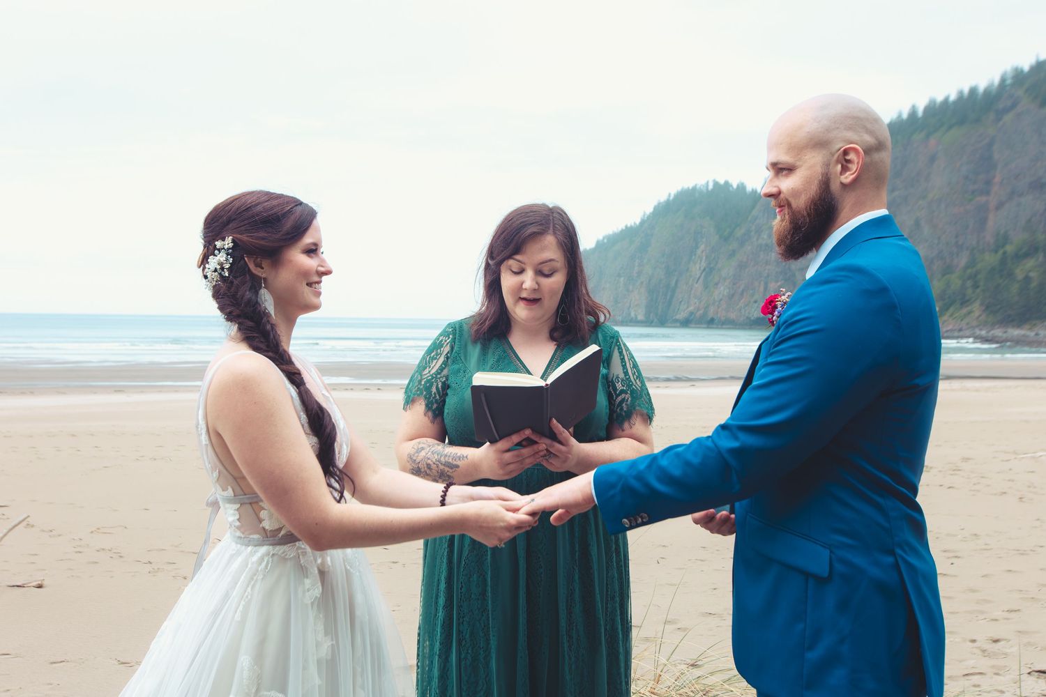 Smiling bride and groom hold hands during beach wedding ceremony while officiant reads from a book.