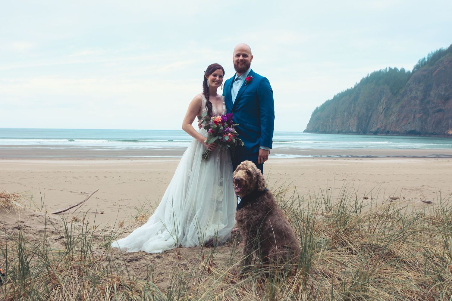 Bride and groom pose with their brown dog on a scenic coastal beach wedding.