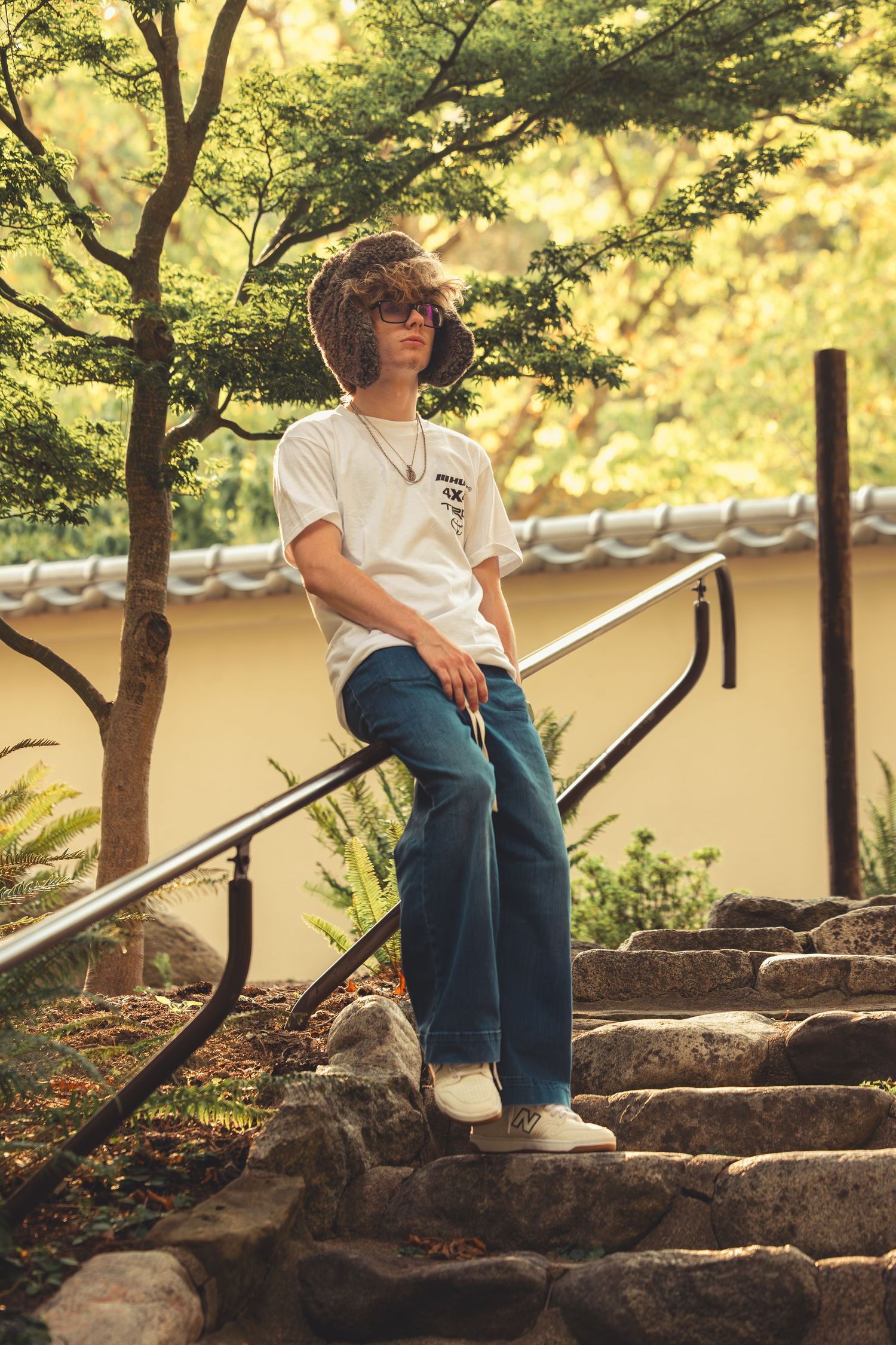Teen in fur hat and white tee leans on metal railing by stone steps in Japanese garden setting.