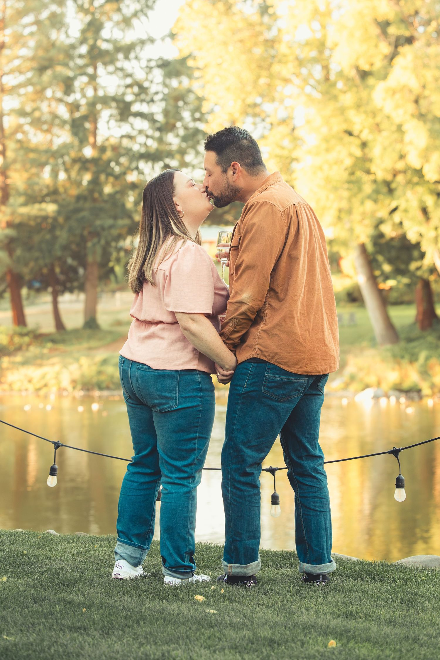 Romantic couple sharing a kiss outdoors by a scenic pond surrounded by golden autumn trees.