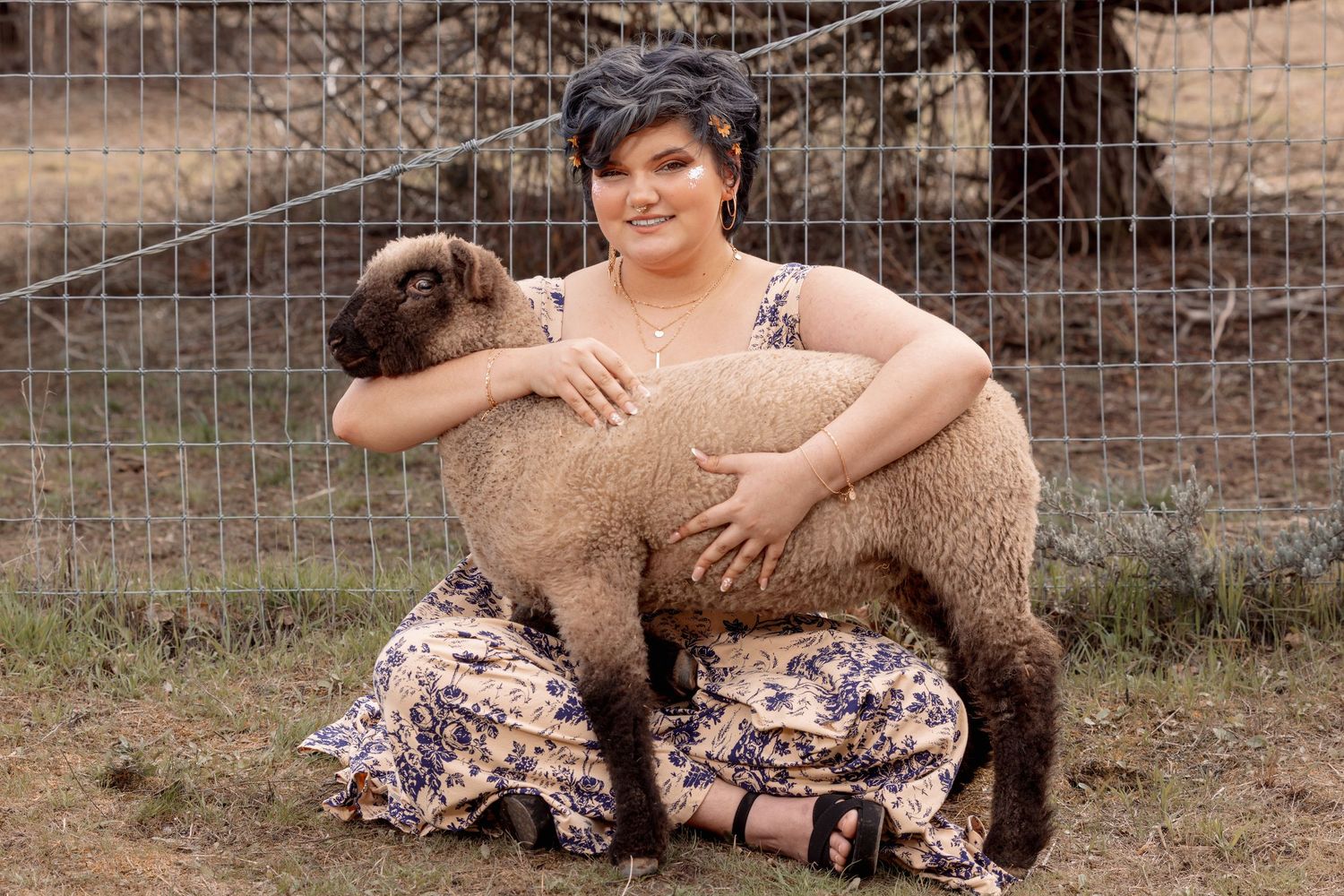 Smiling woman in floral dress sitting outdoors on grass, embracing a fluffy brown lamb.