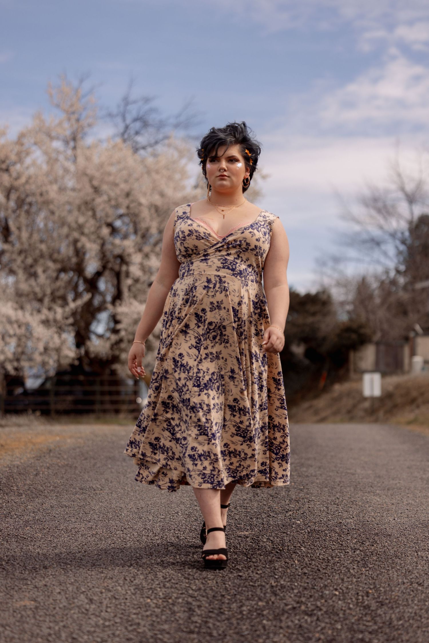 Woman in floral midi dress and black heels walking on road beside white flowering spring tree.