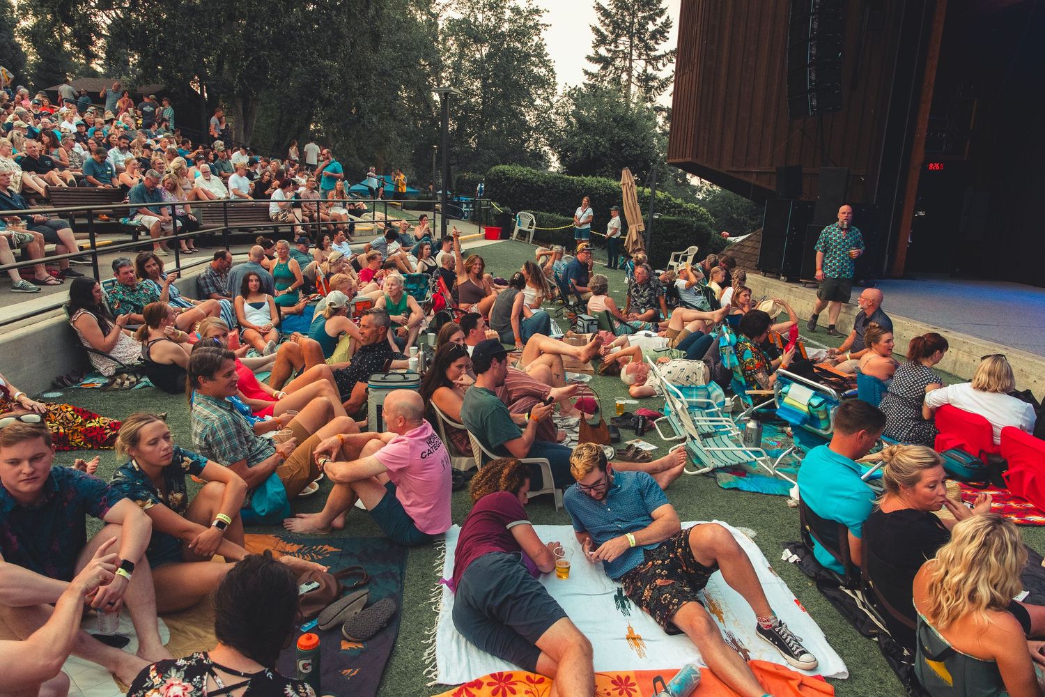 Crowd of concert-goers sitting on blankets and chairs at an outdoor amphitheater during a summer music event.