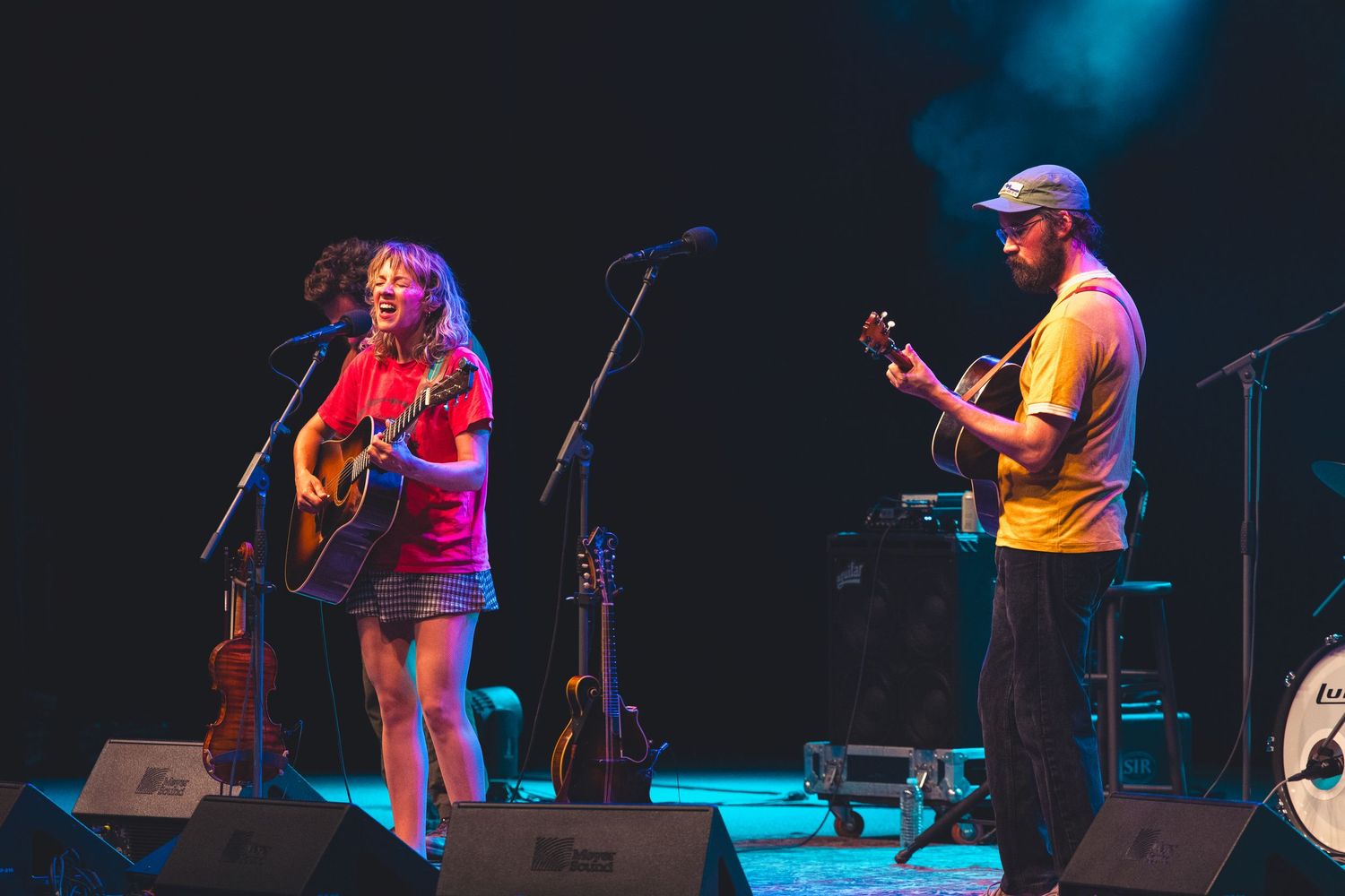 Female guitarist and male ukulele player performing together under colorful stage lights at a live concert.