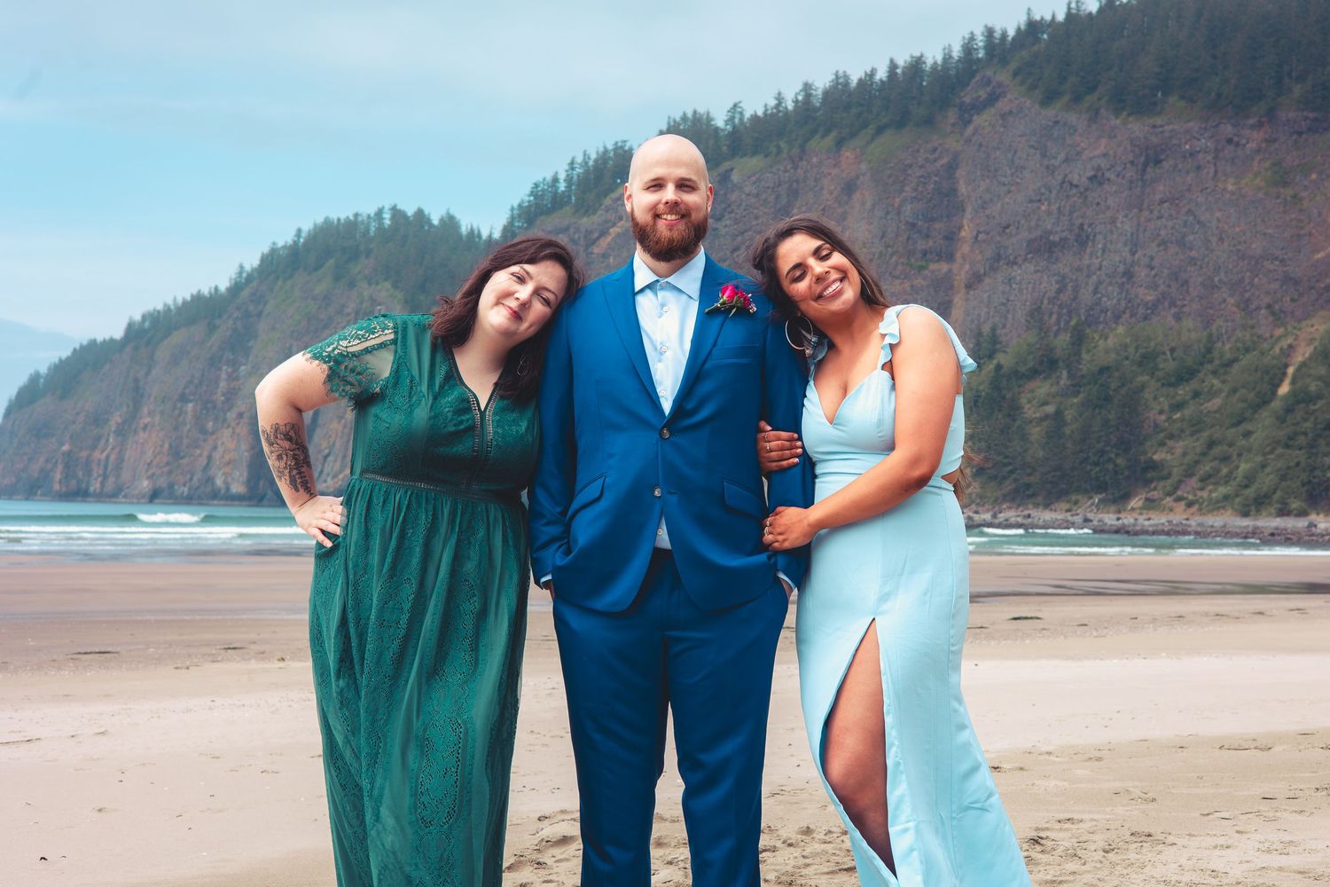 Groom in blue suit smiles with two bridesmaids in green and light blue gowns on a beach.