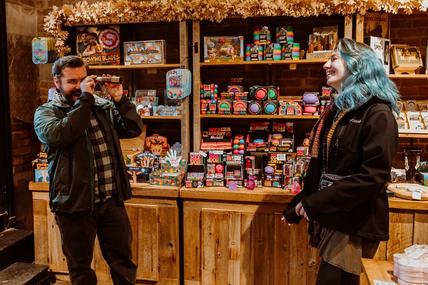 Bearded man photographing a smiling blue-haired woman in a festive shop.