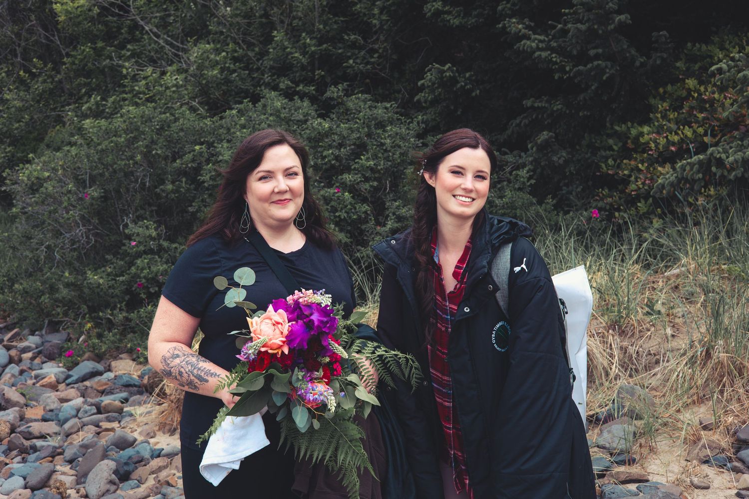 Two women smiling outdoors on a rocky beach, one holding a vibrant colorful floral bouquet.