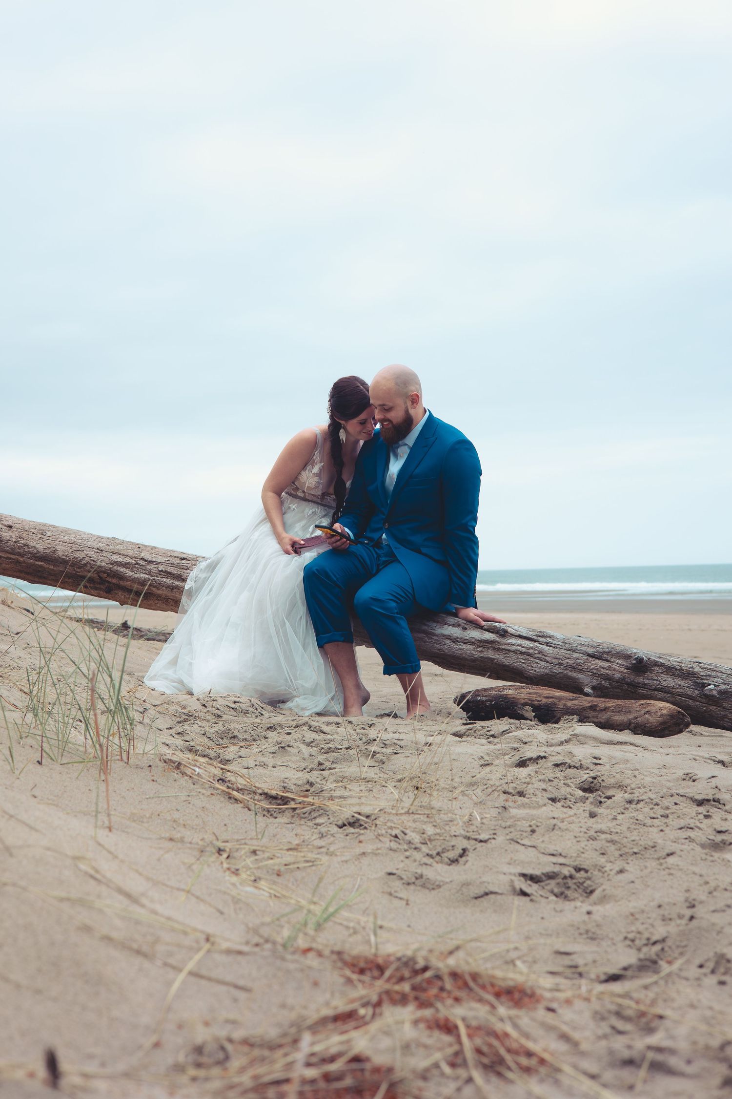 Bride and groom share an intimate moment leaning close together on driftwood logs at a cloudy beach.