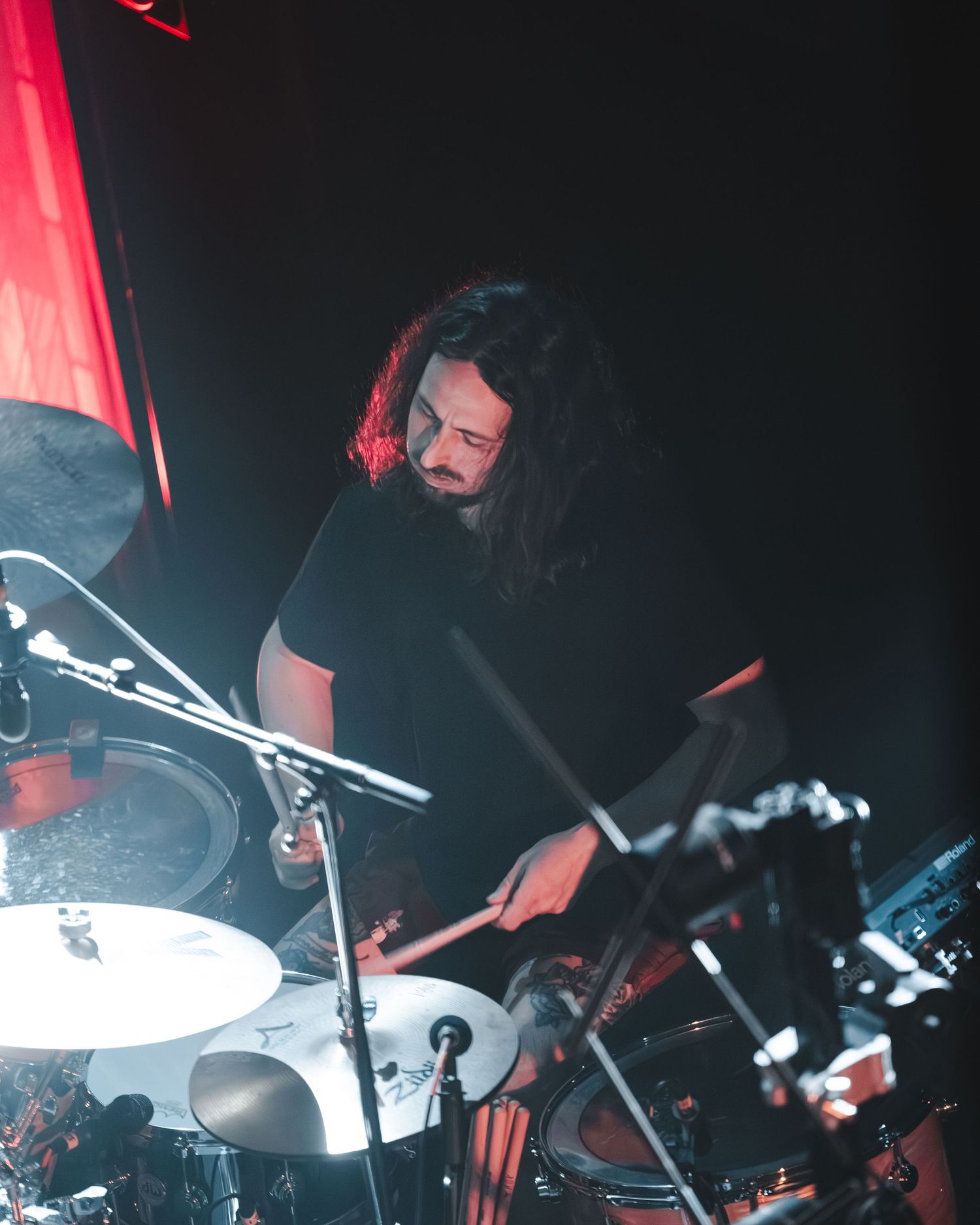 Long-haired drummer playing drums under red stage lighting during a live music performance.
