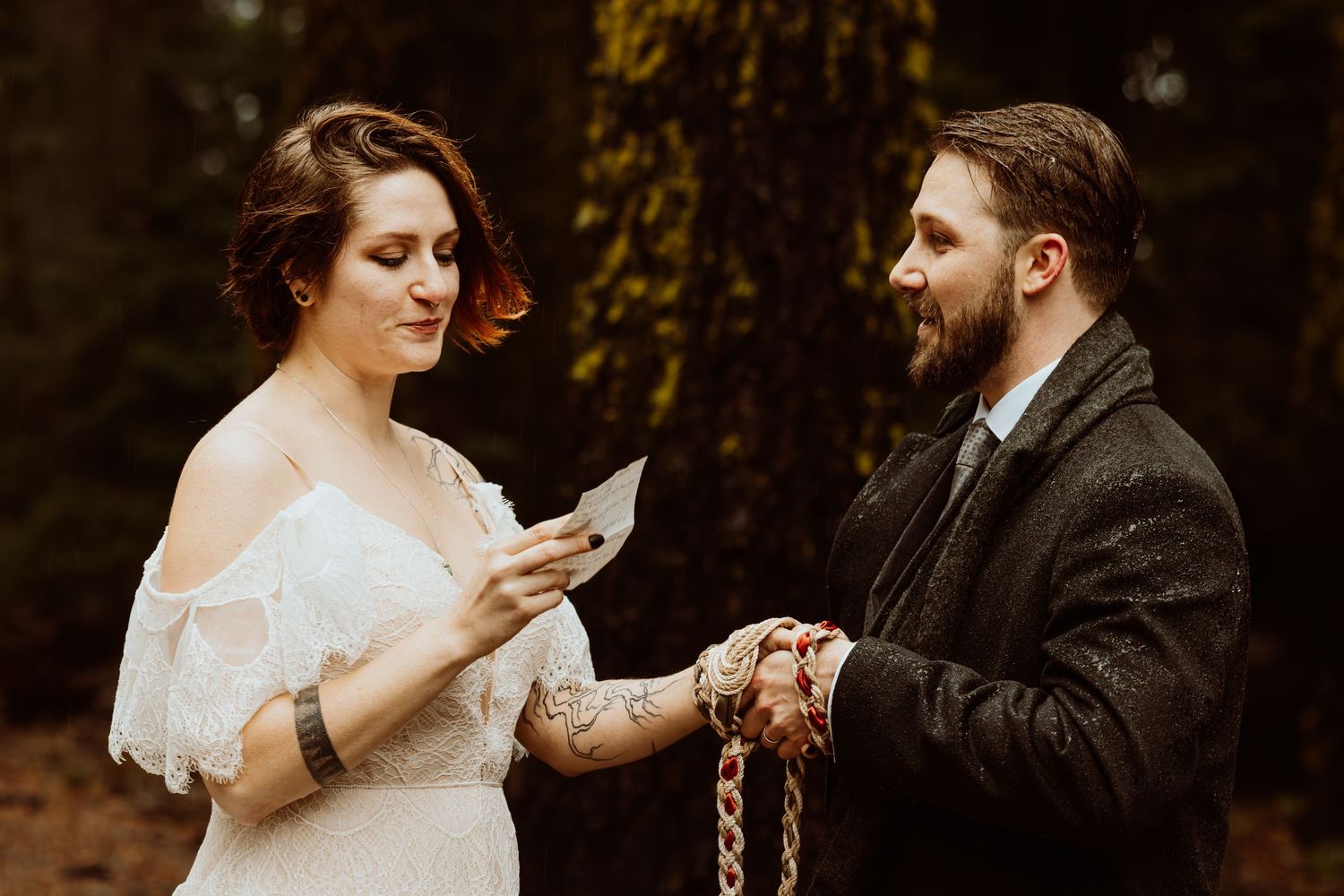 Bride reads wedding vows while groom holds her hands with a rope handfasting ceremony in a forest.