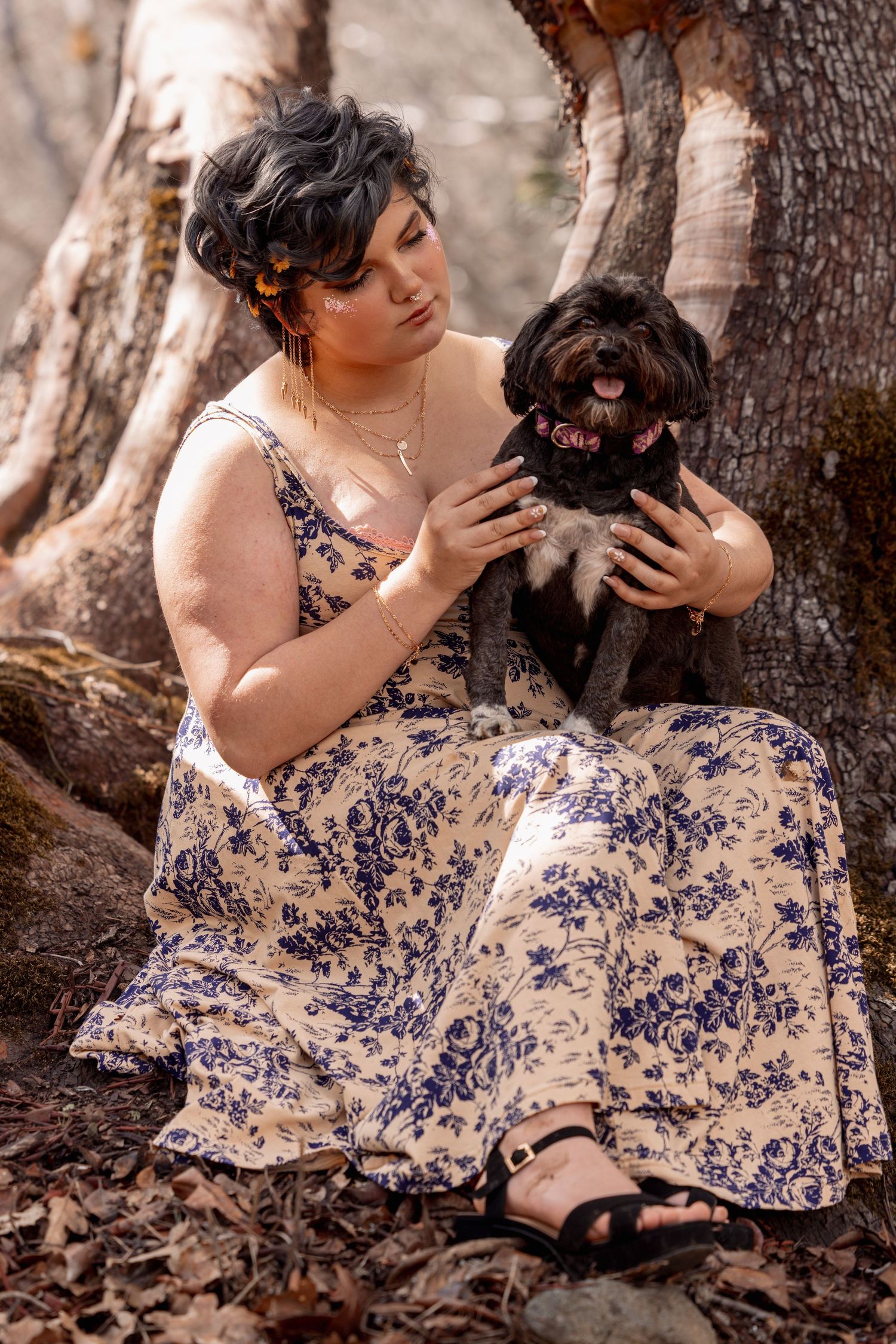 Woman in cream floral dress sitting under tree, holding small black dog on her lap.