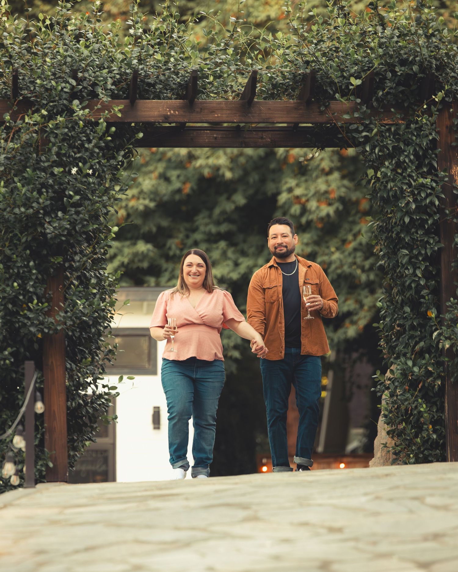 Couple walking hand in hand through a vine-covered pergola archway holding drinks outdoors.