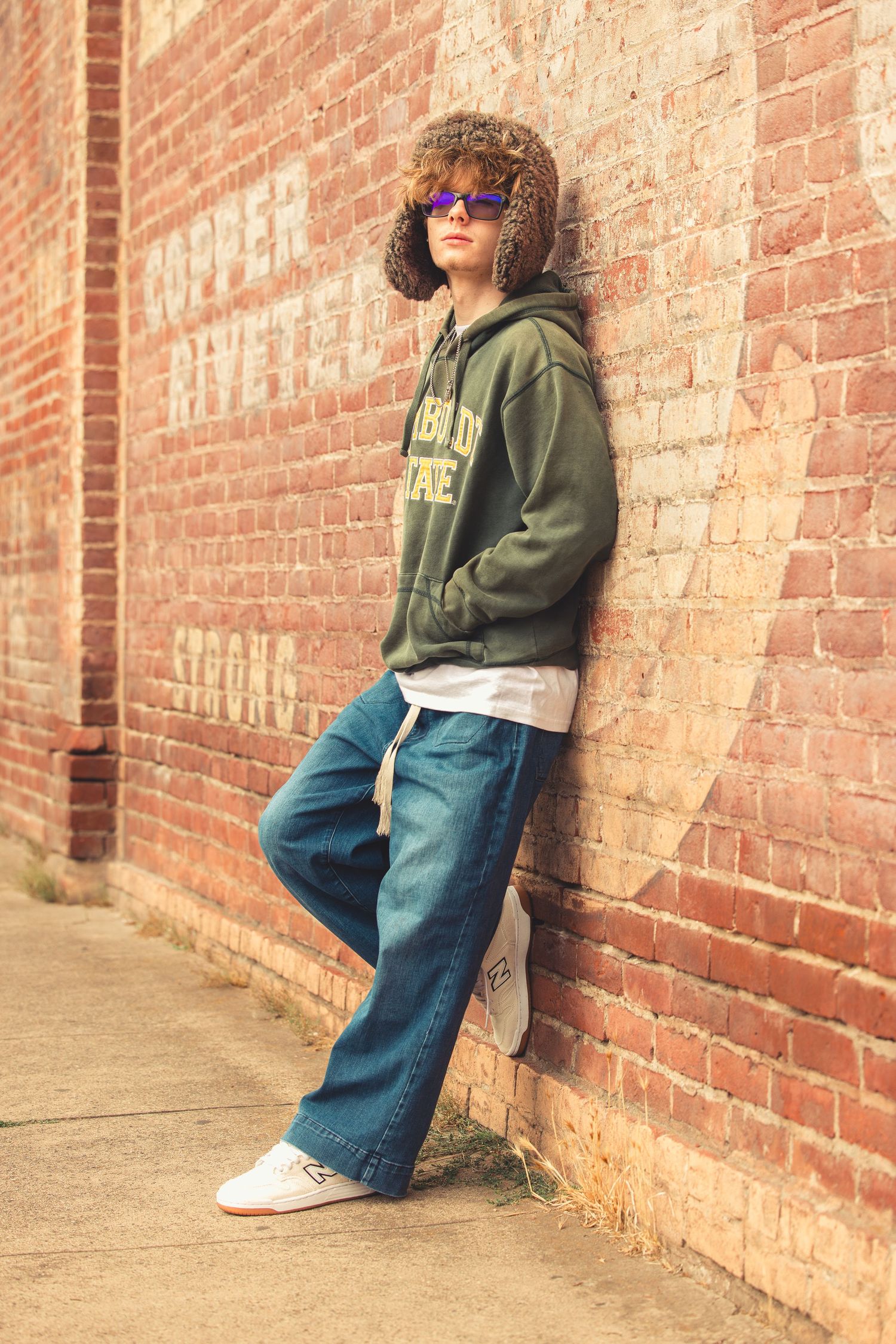 Teen in green Humboldt State hoodie, fur hat, and wide-leg jeans leans against vintage brick wall.