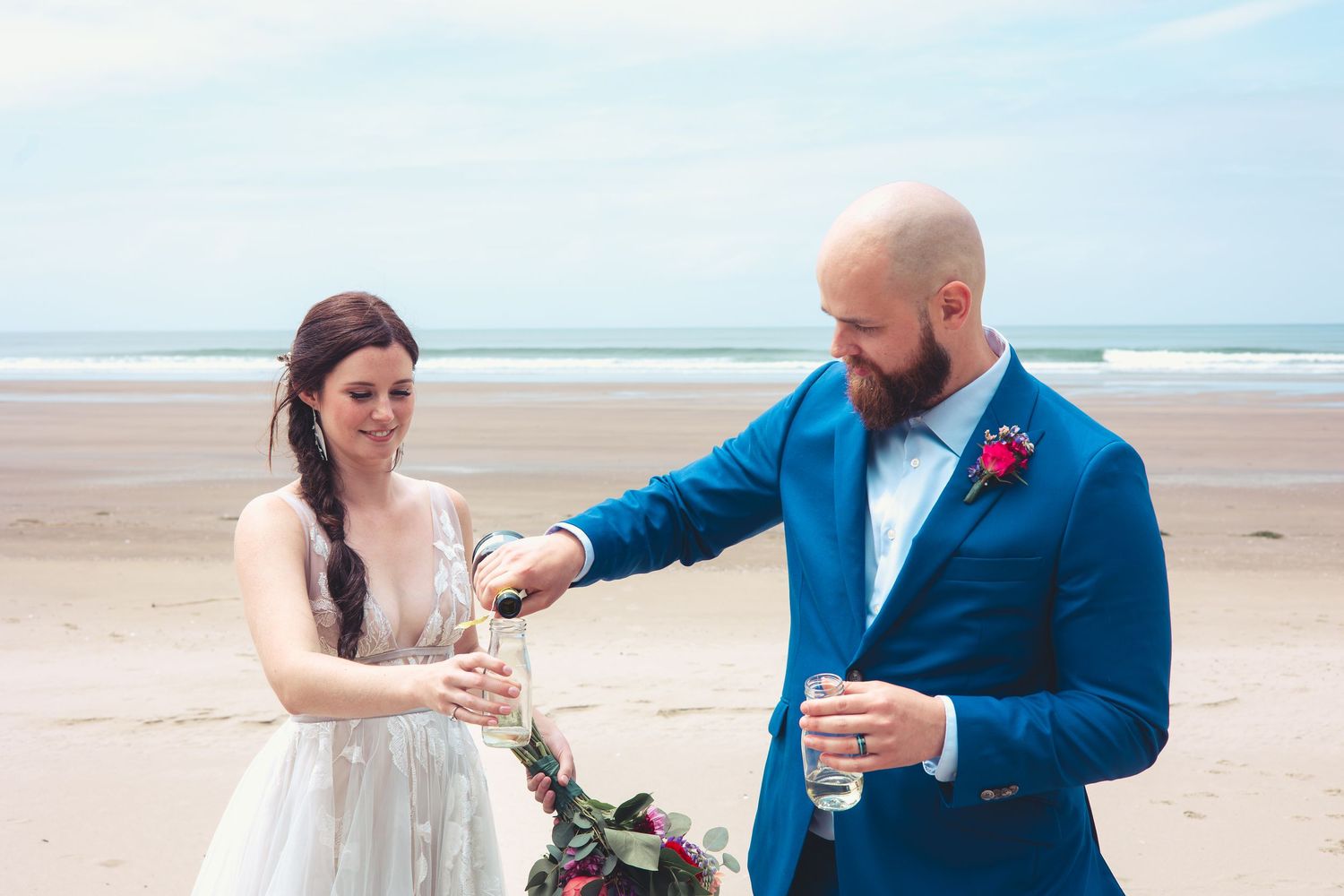 Groom pouring champagne for bride on a sandy beach during their romantic seaside wedding celebration.