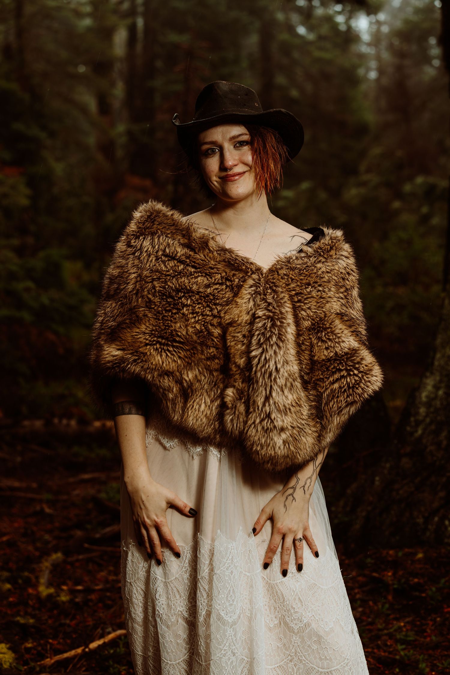 Bride in white lace wedding dress and fur stole wearing black hat, posing in misty forest setting.