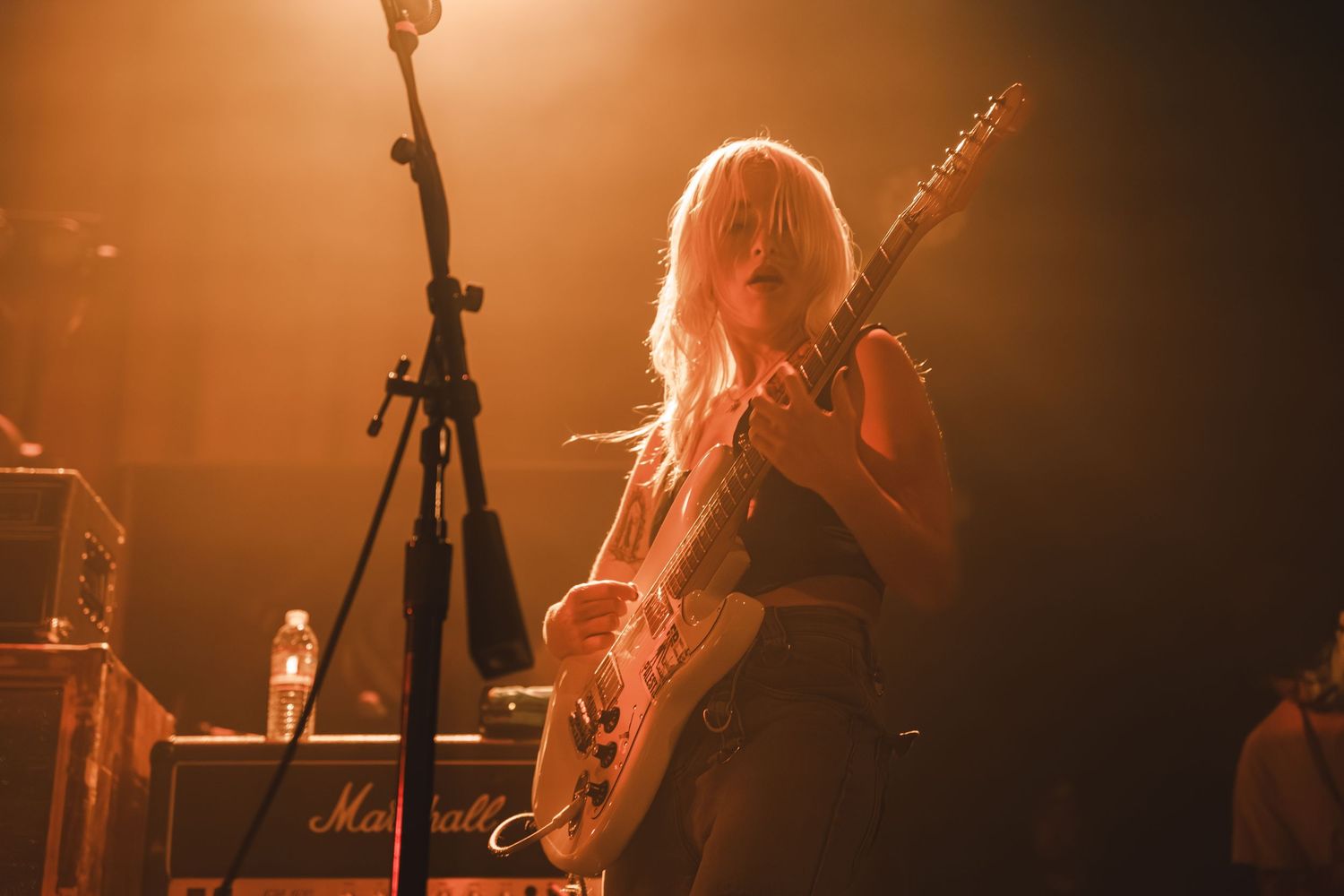 Blonde female guitarist performs on stage with electric guitar under warm orange stage lighting.