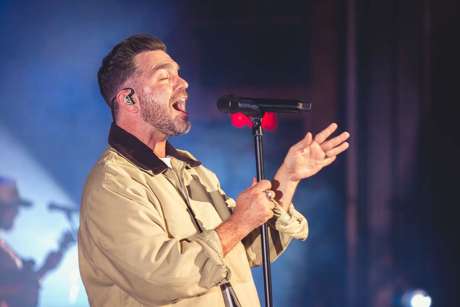 A male singer passionately performs at a microphone stand under warm stage lighting.