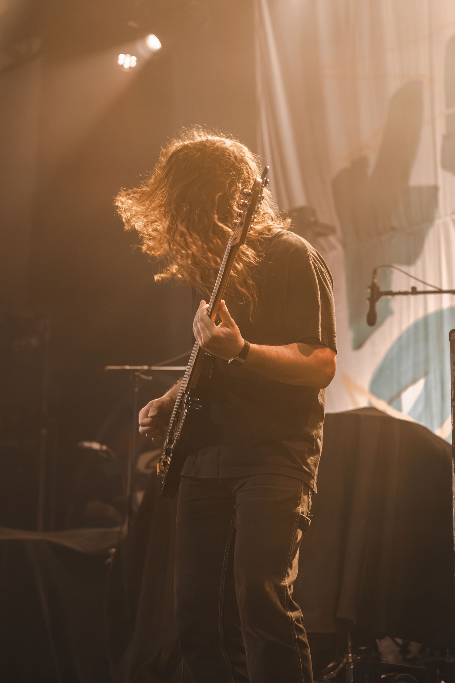 Long-haired guitarist performing on stage with back turned, bathed in warm golden stage lighting.