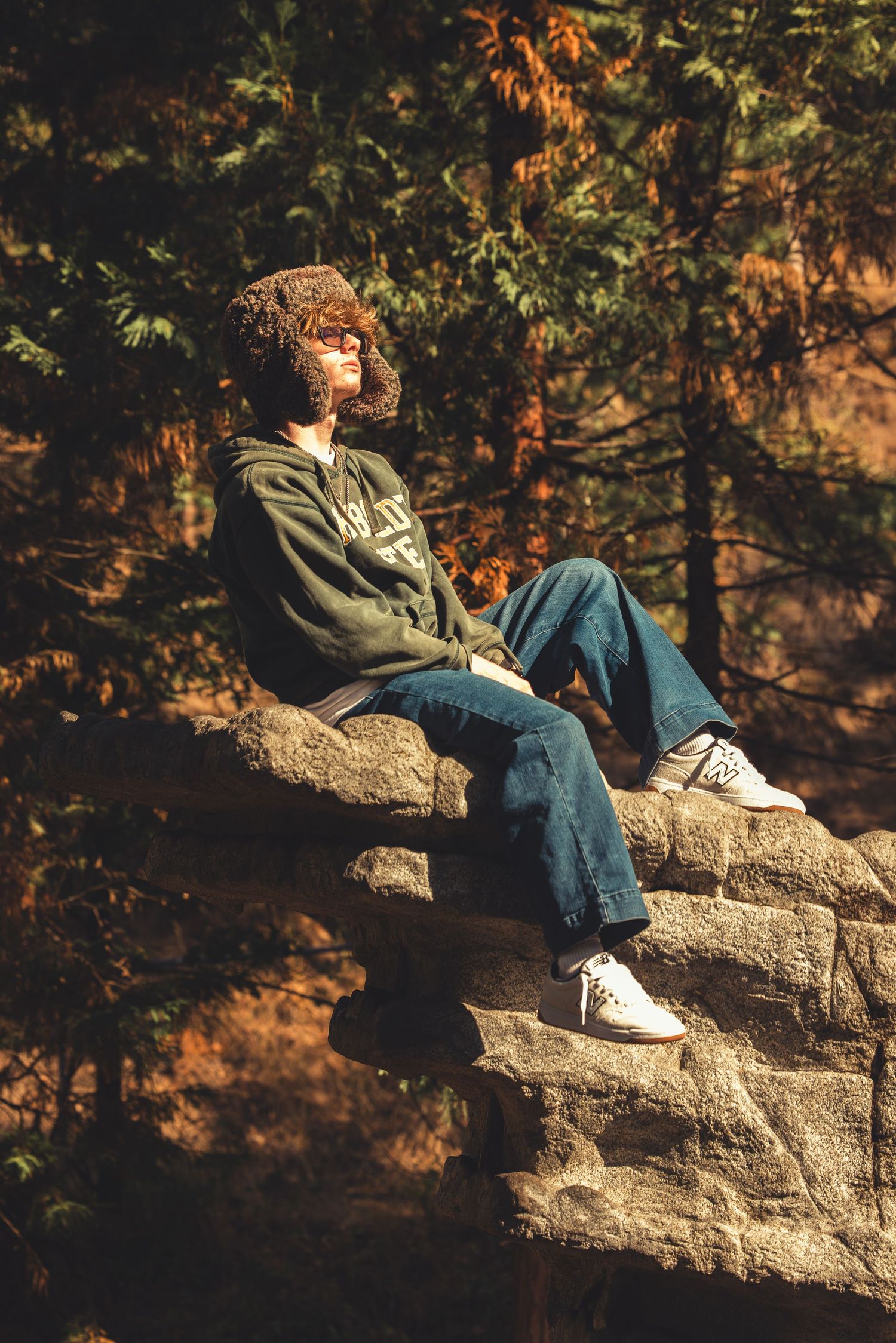 Teen in fur hat and green hoodie reclines on stone ledge surrounded by autumn forest foliage.