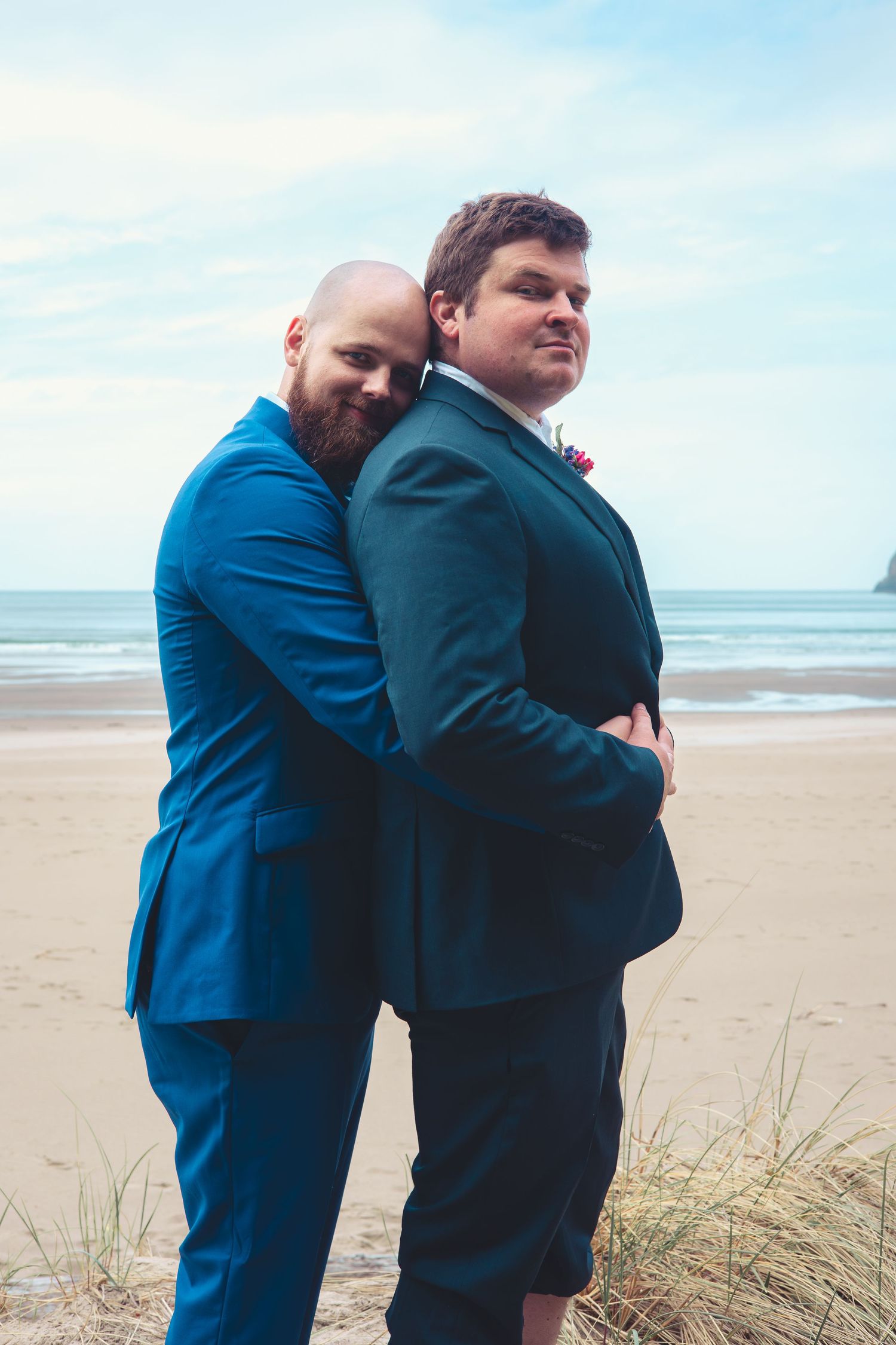 Two grooms in blue suits share a warm embrace on a sandy beach wedding.
