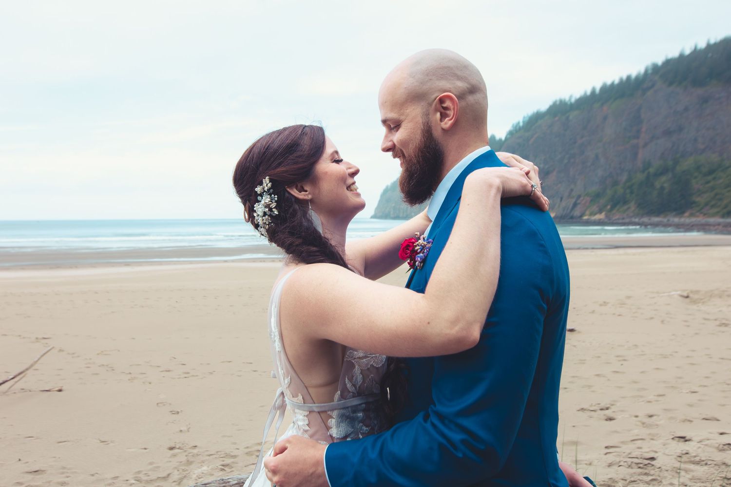 Laughing newlyweds embrace joyfully on a sandy beach with a scenic forested hillside backdrop.