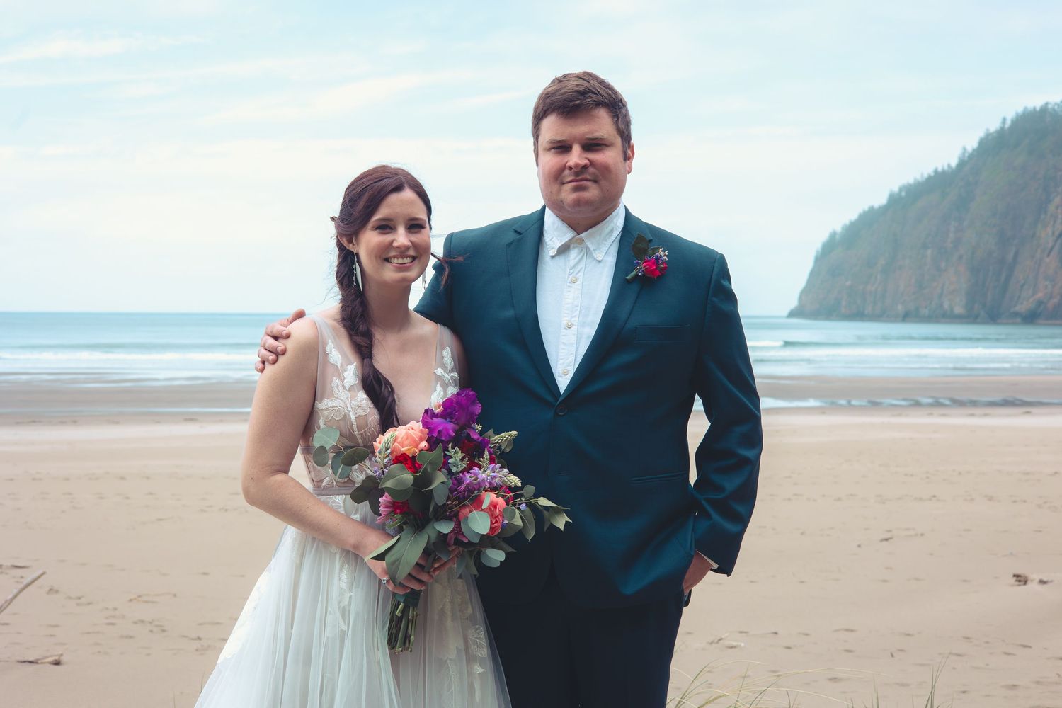 A bride holding a colorful bouquet and groom in a navy suit smile together on a sandy beach.