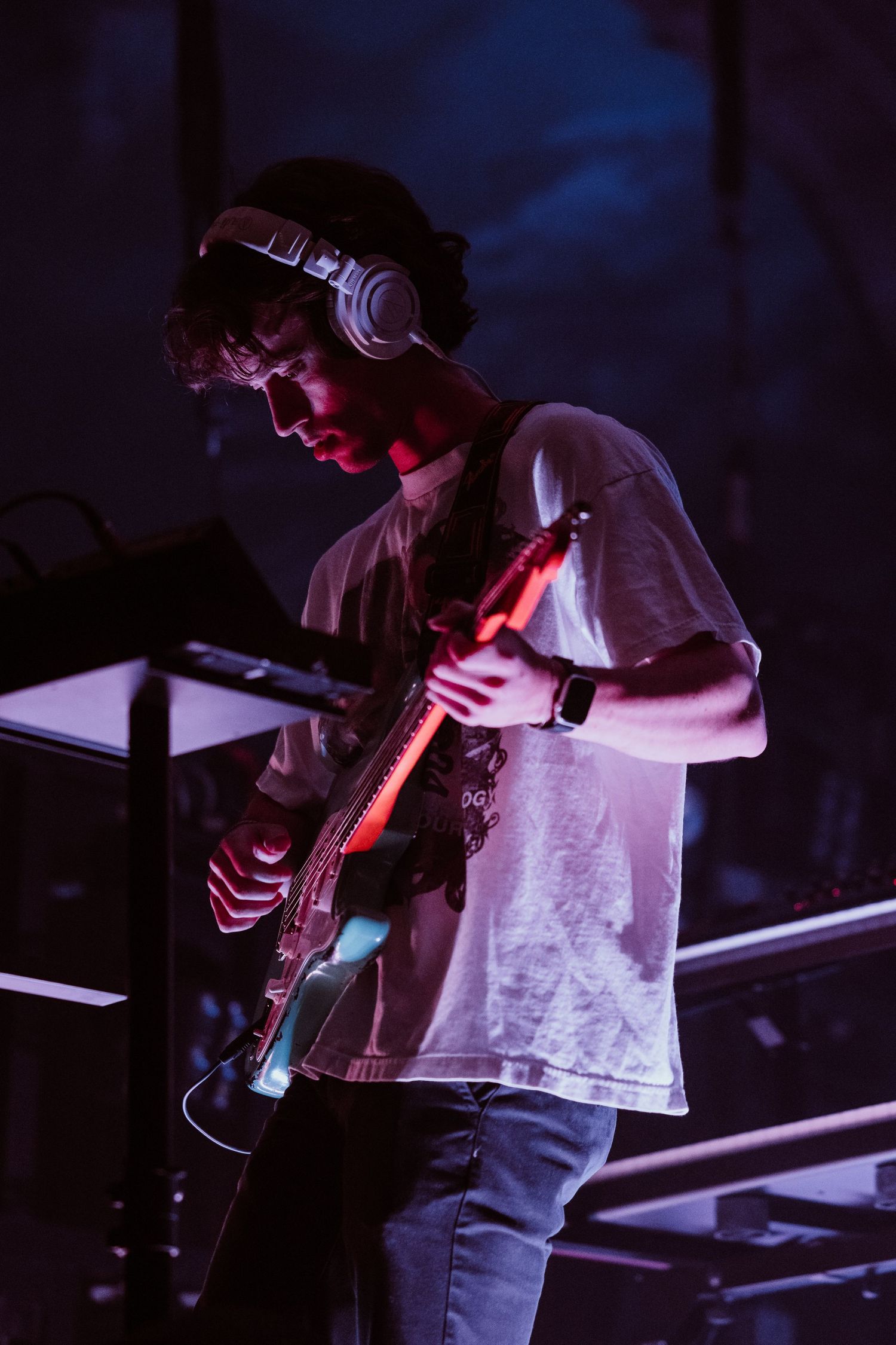Young male guitarist wearing headphones performing under purple and red stage lights.