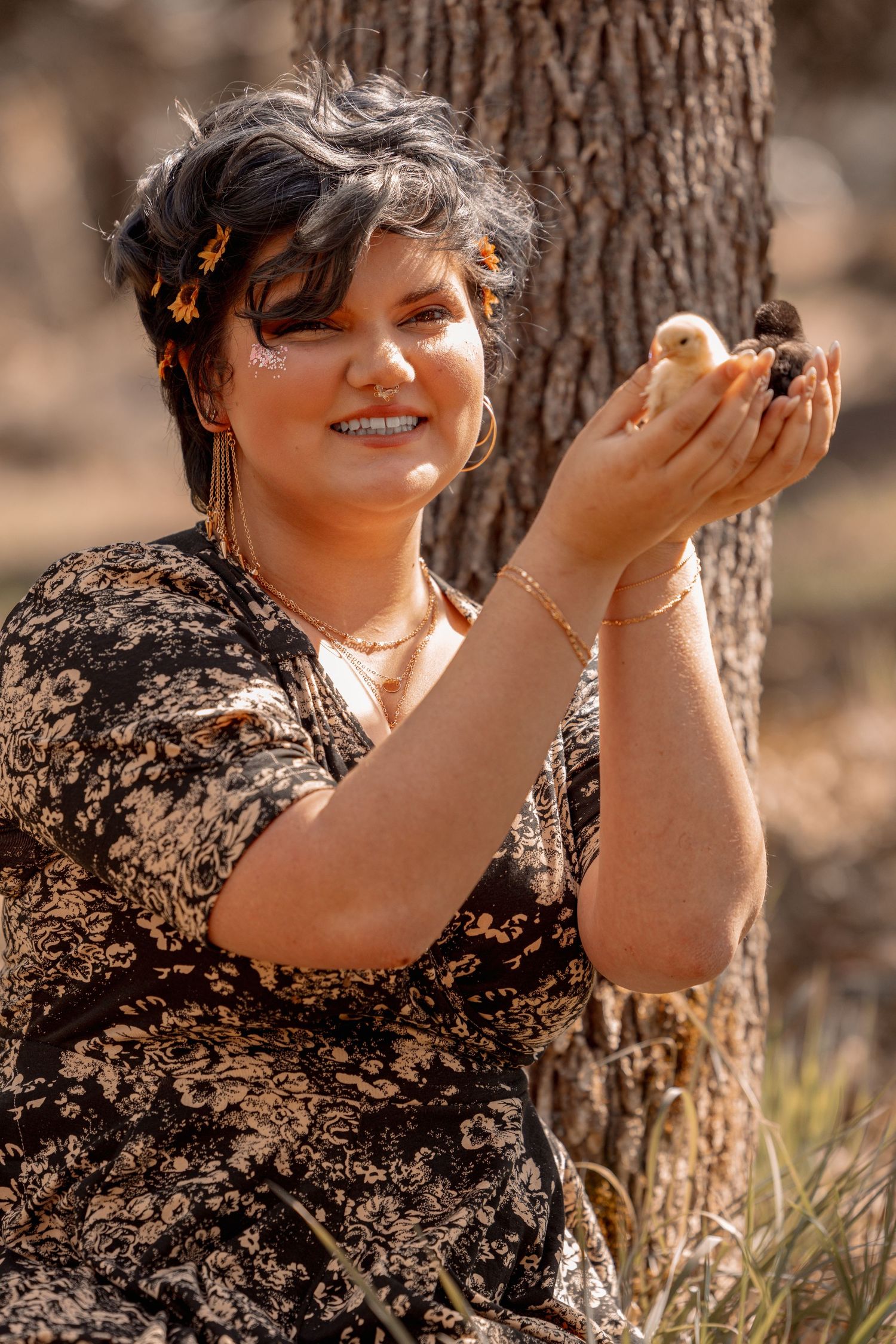 Smiling woman with sunflower hair accessories holds two baby chicks in her hands outdoors.