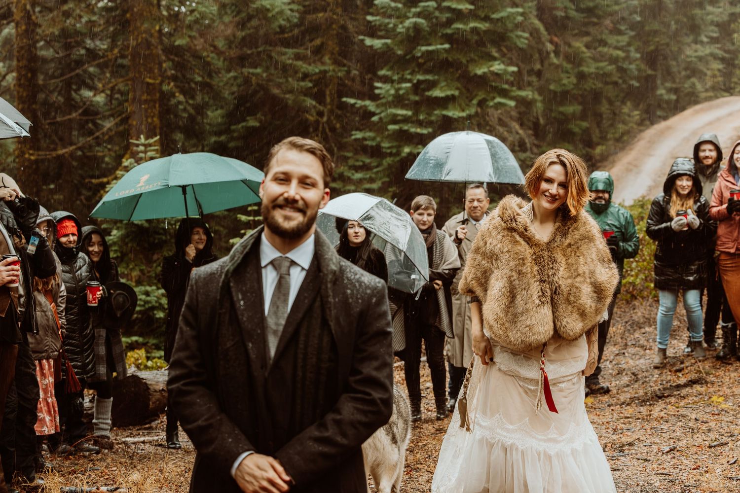 Couple shares joyful moment during outdoor rainy forest wedding ceremony with guests holding umbrellas.