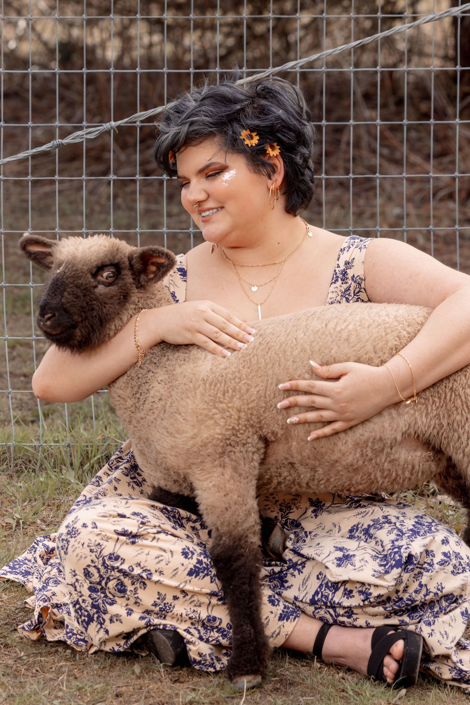 Woman in floral dress sitting on ground hugging a brown lamb near wire fence.