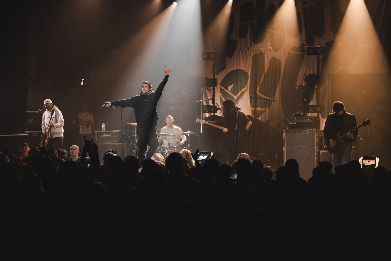 Band performing energetically on a dramatic stage with spotlights and crowd silhouettes at a live rock concert.