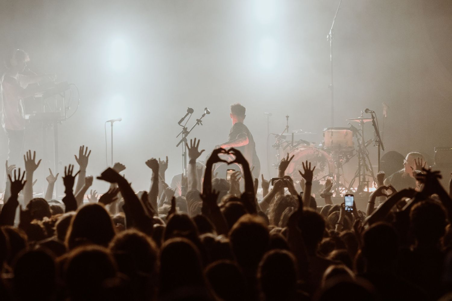 Concert crowd raises hands toward solo performer on brightly lit stage.