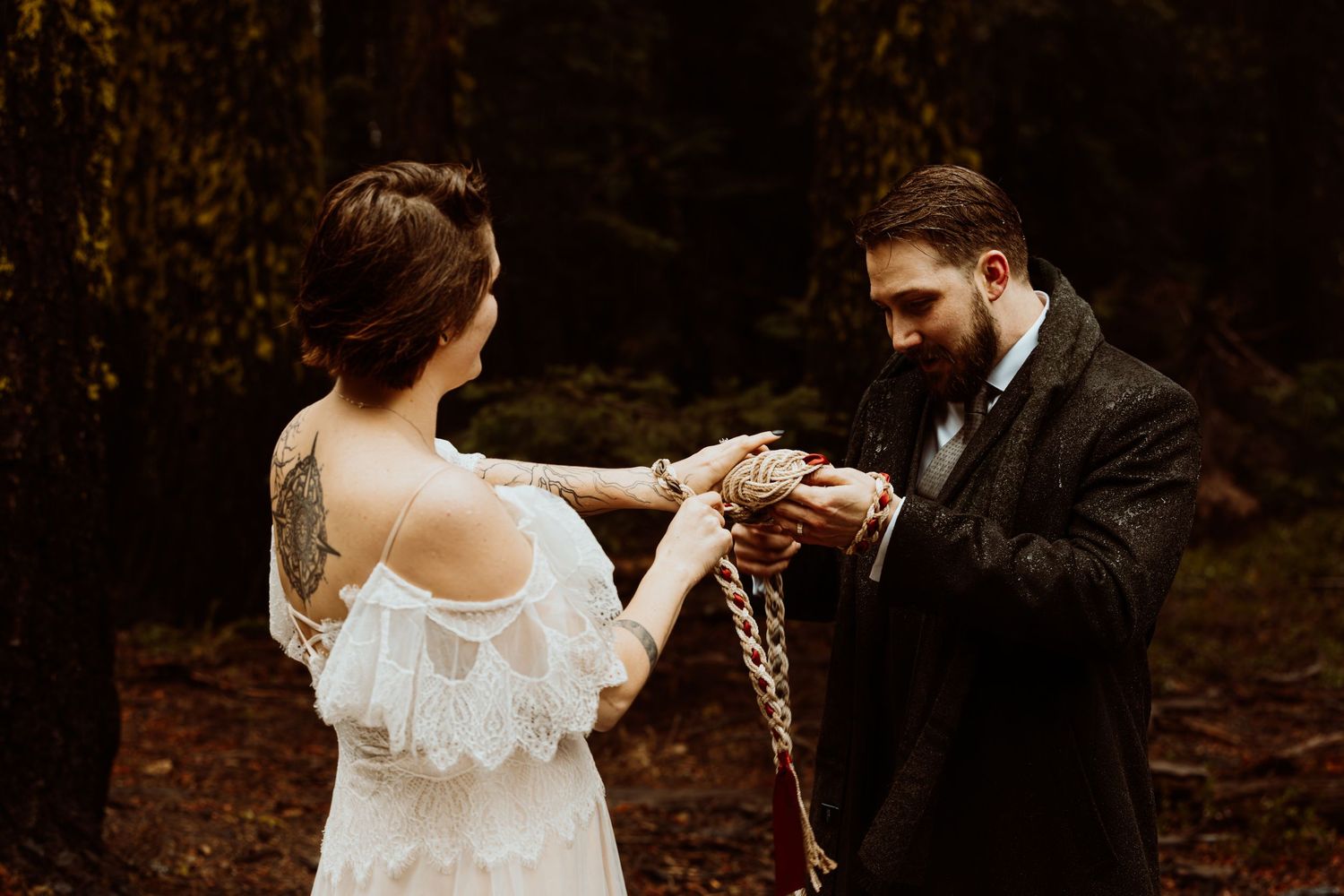 Couple performing a handfasting ceremony with a braided cord in a moody forest setting.