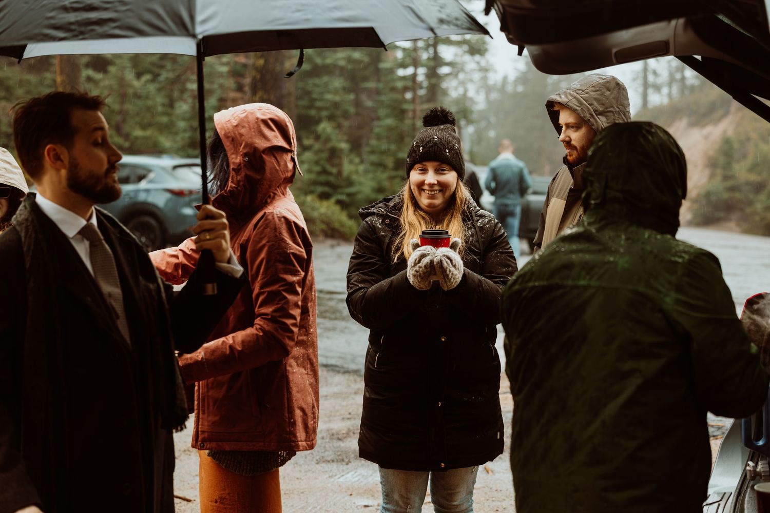 Wedding guests chat under umbrellas in the rain at an outdoor forest ceremony location.