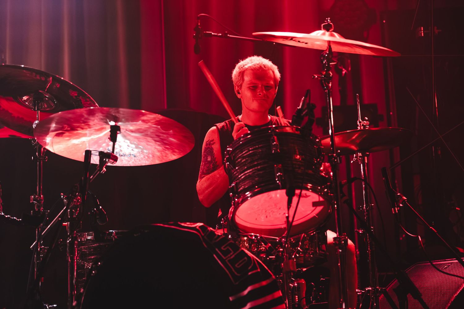 Drummer performing on stage under dramatic red lighting, focused on playing full drum kit at live concert.