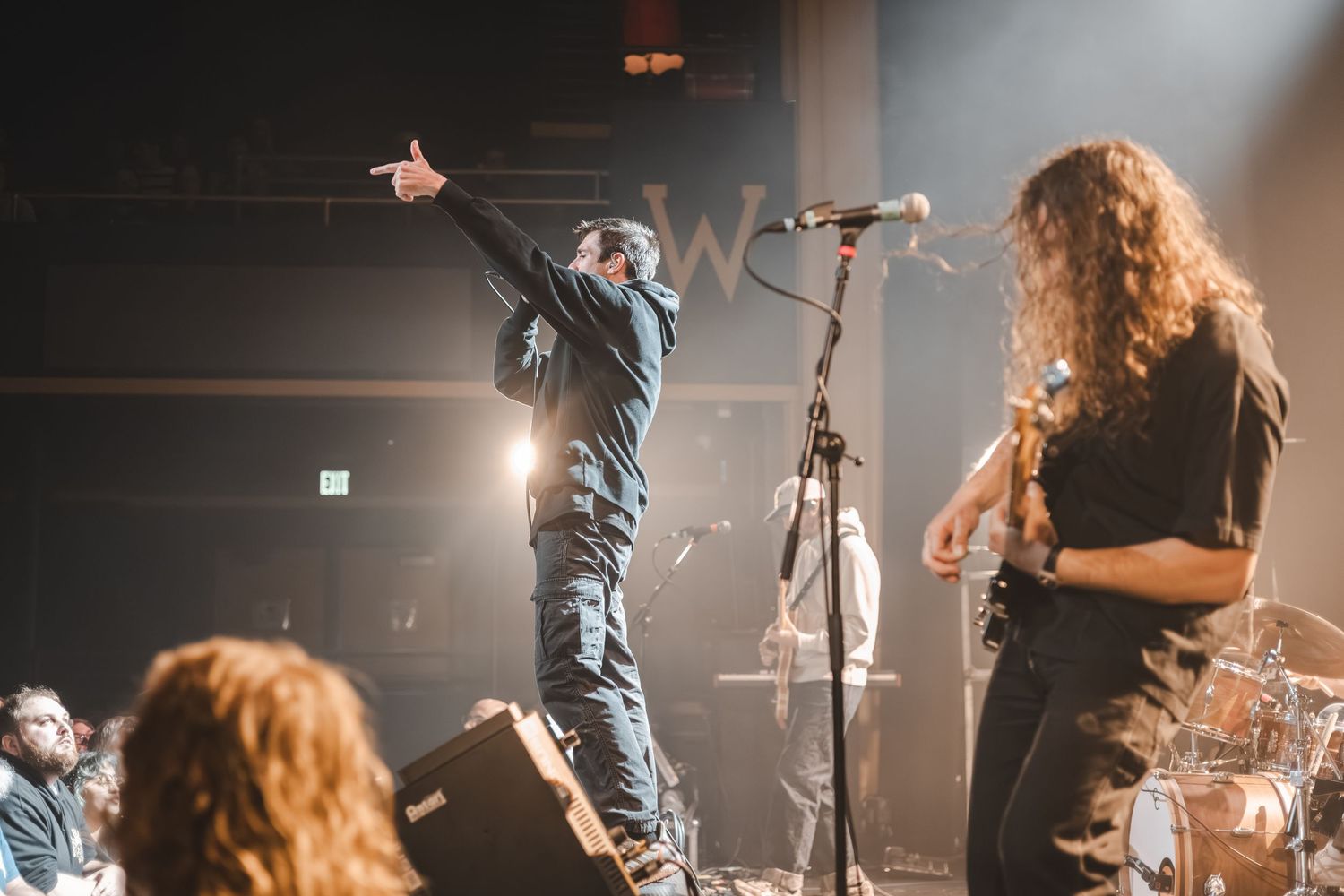 Rock band performing live on stage with vocalist gesturing to crowd and guitarist playing under dramatic stage lighting.