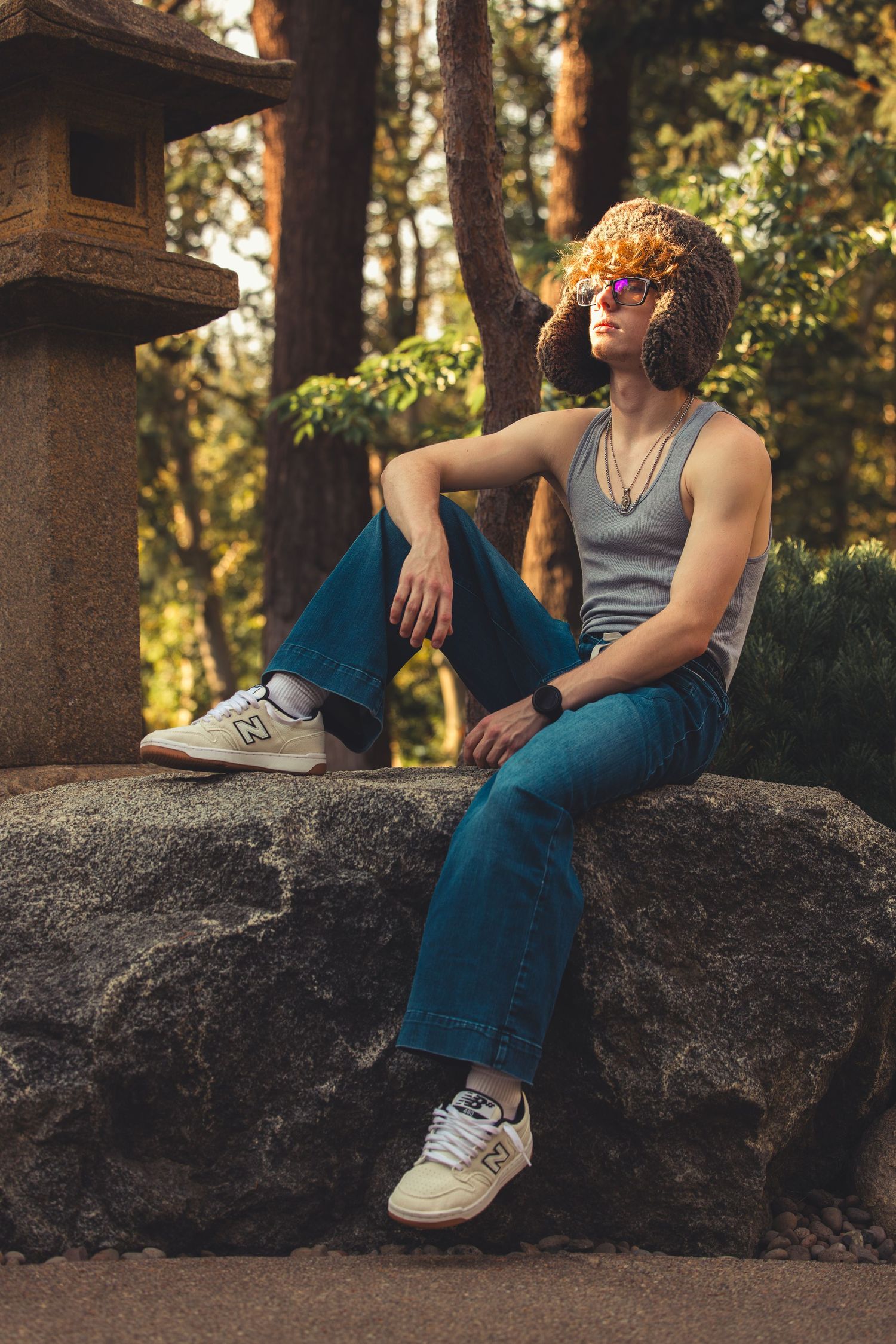 Young man in gray tank top, jeans, and New Balance sneakers sitting on rocks in a Japanese garden.