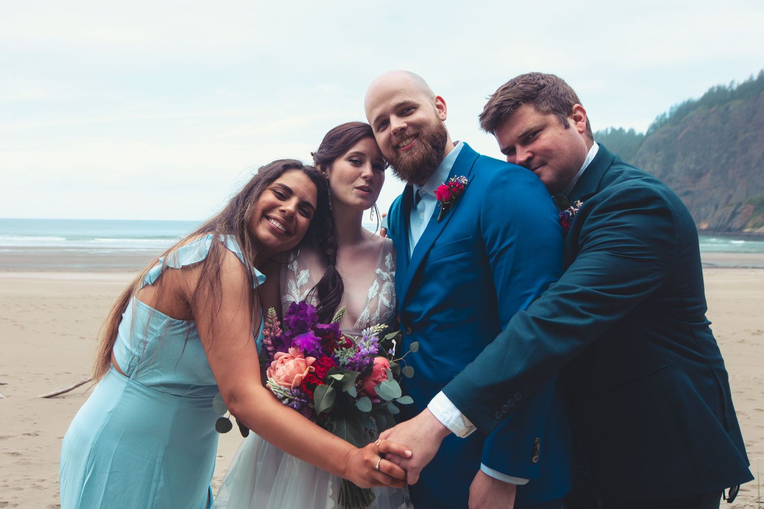 Wedding party of four smiling and holding hands on a scenic beach celebration.