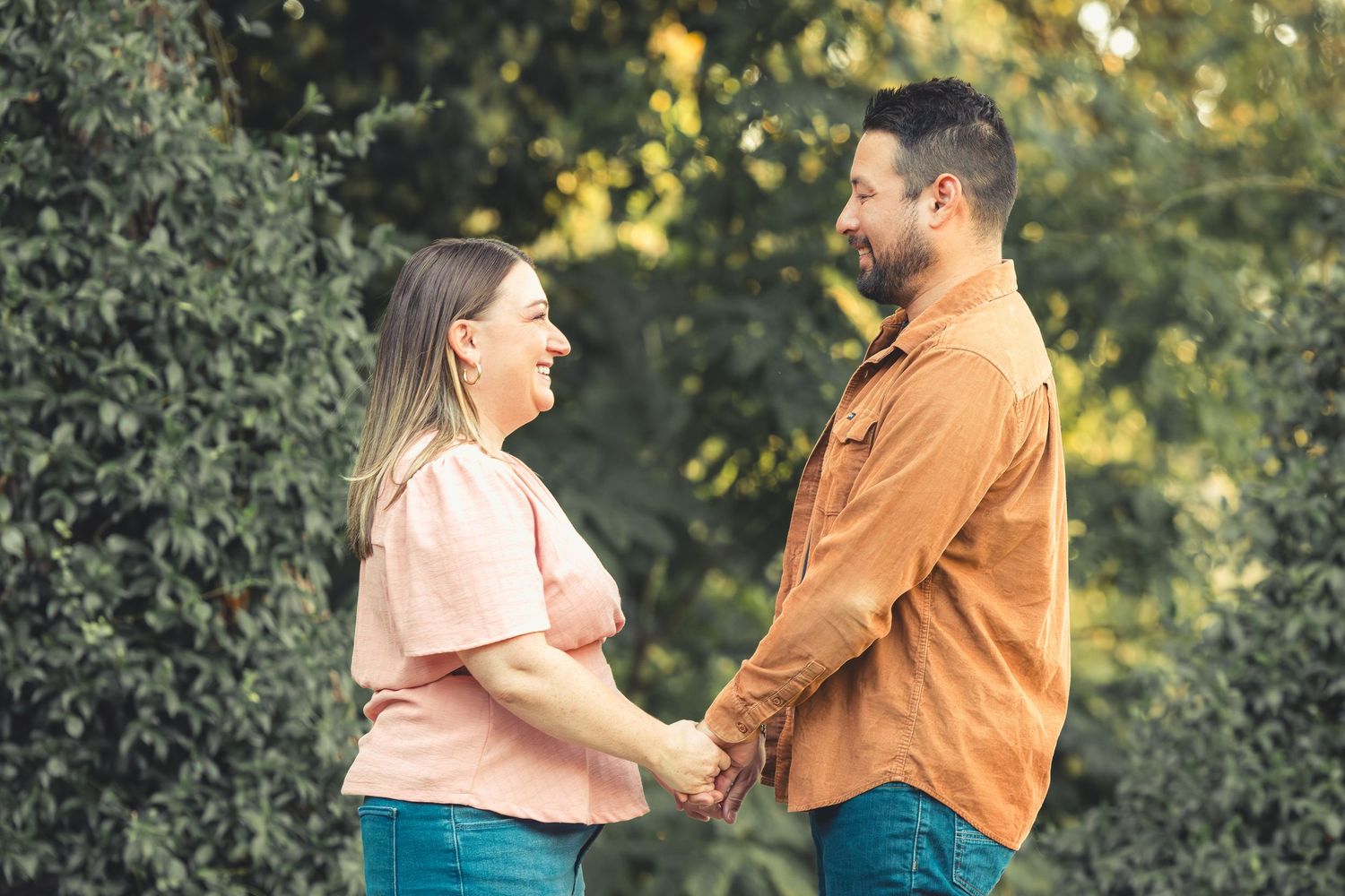 Couple holding hands and smiling at each other in a lush green garden setting on a sunny day.