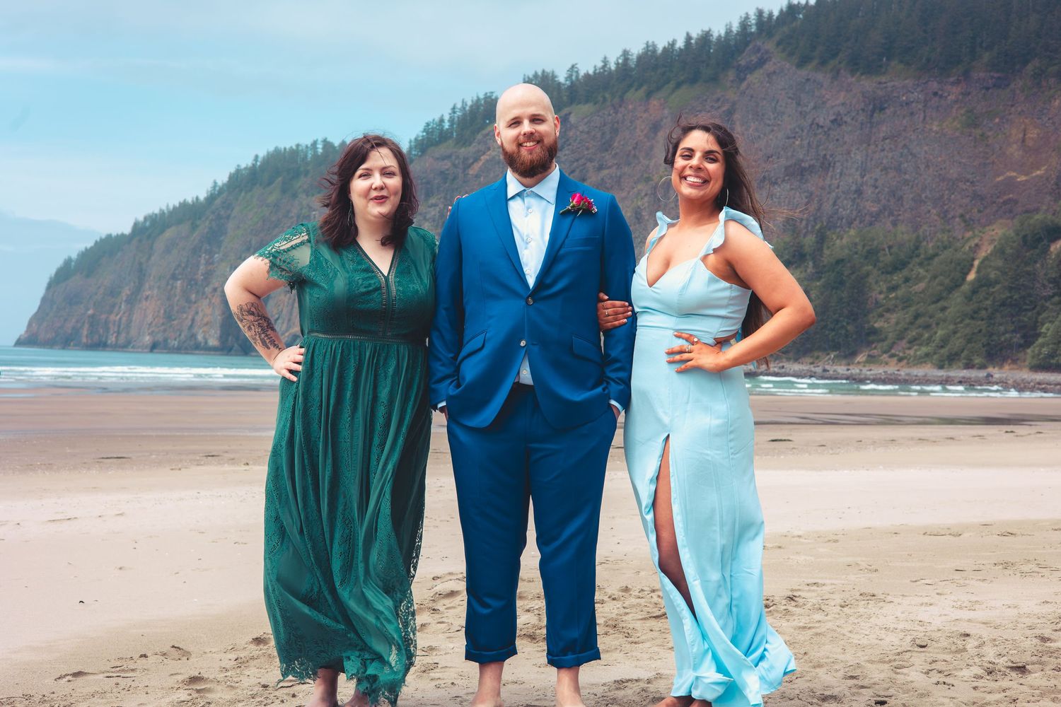 Groom in blue suit posing with two bridesmaids in green and light blue dresses on a beach.