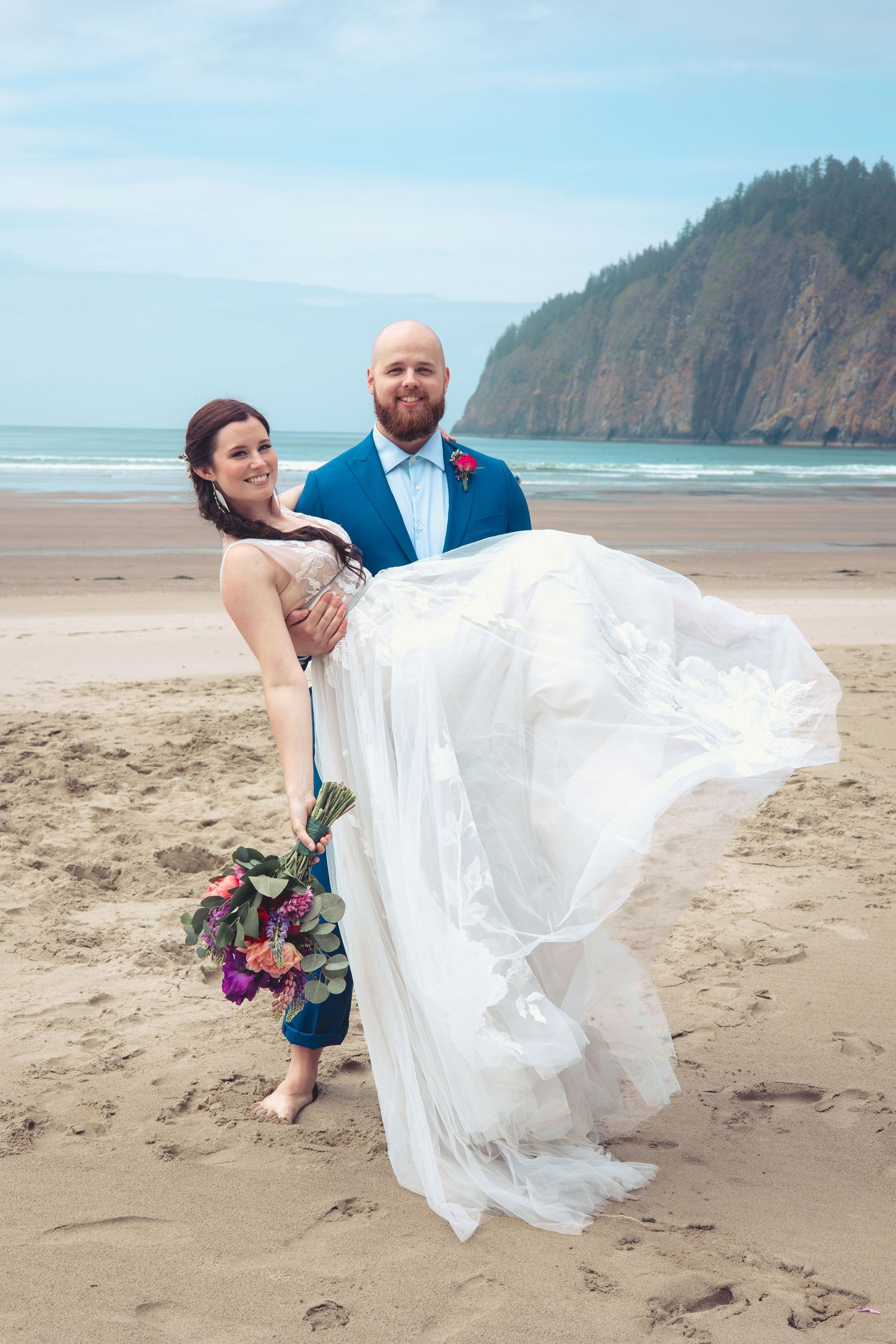 Bride and groom pose barefoot on sandy beach, her white gown flowing in wind, rocky cliffs in background.