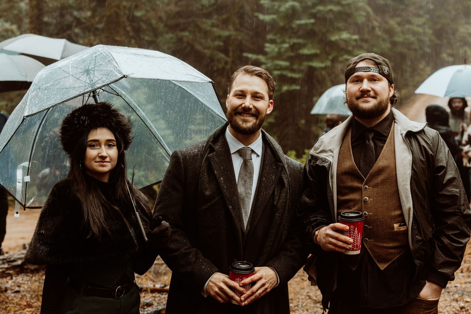 Three wedding guests smile under clear umbrellas in rainy forest wedding setting.