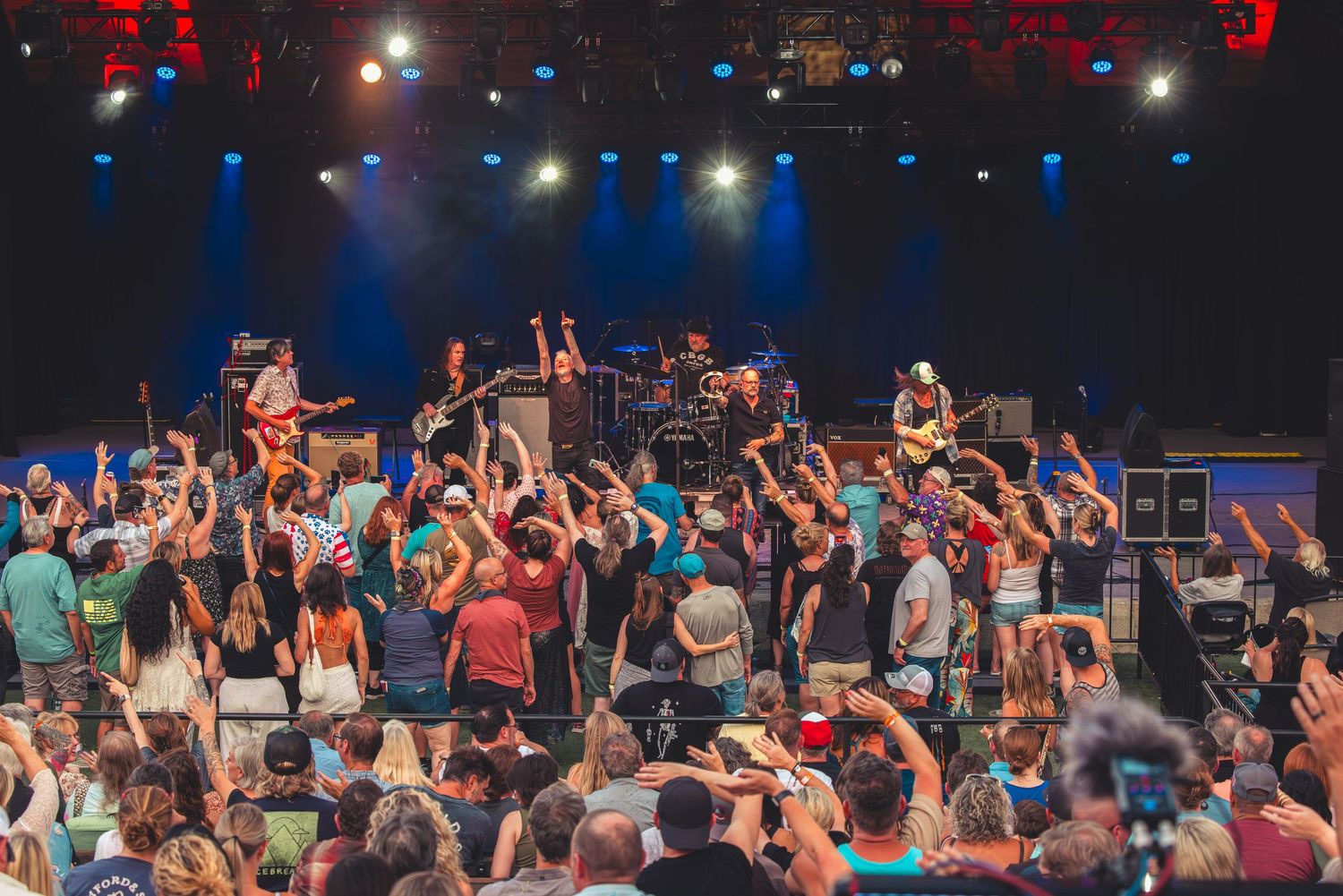 Rock band performing on stage as an enthusiastic crowd raises their hands.