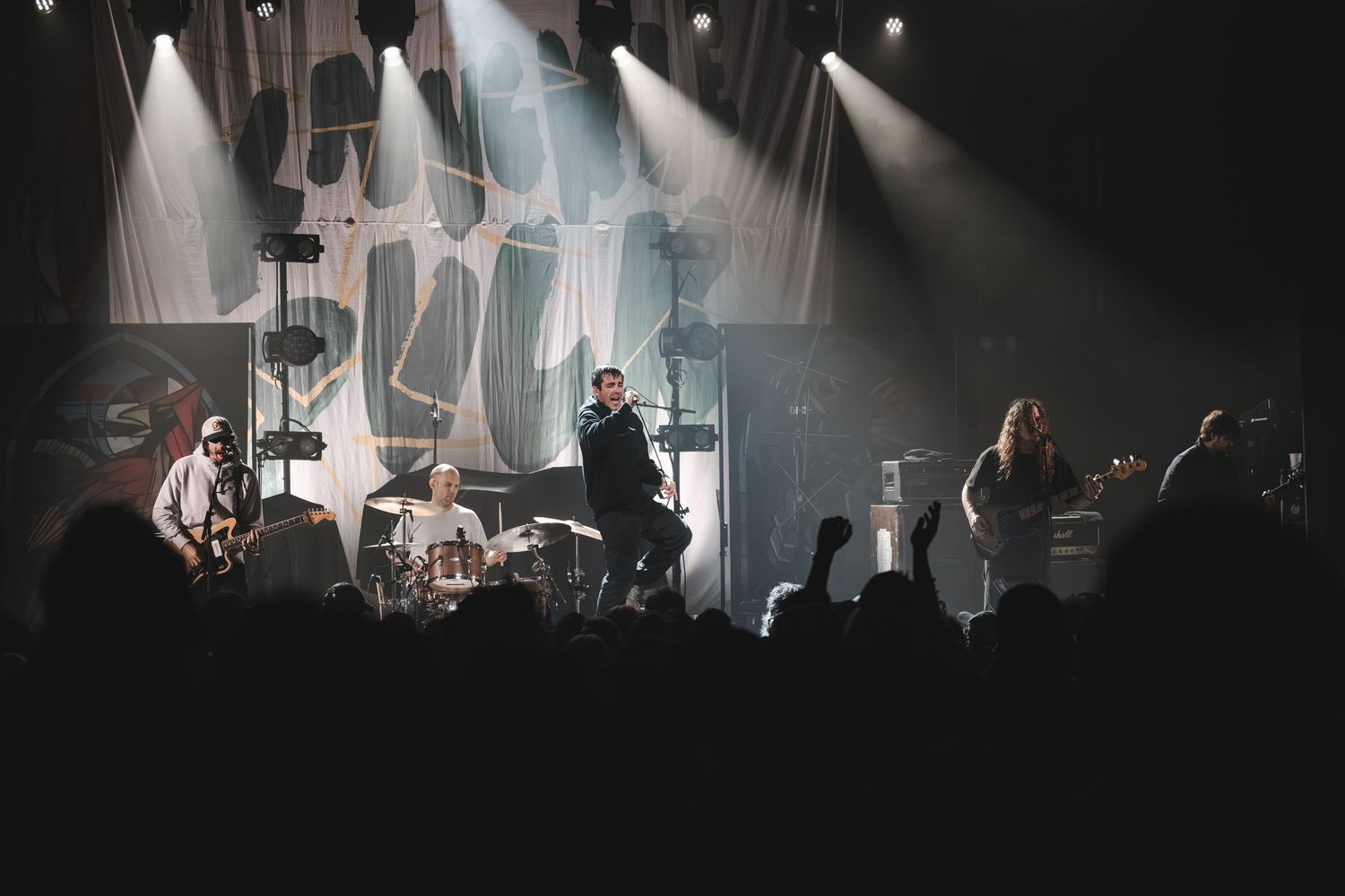 Full band performing under white stage lights with abstract backdrop and crowd silhouettes at indoor music venue.